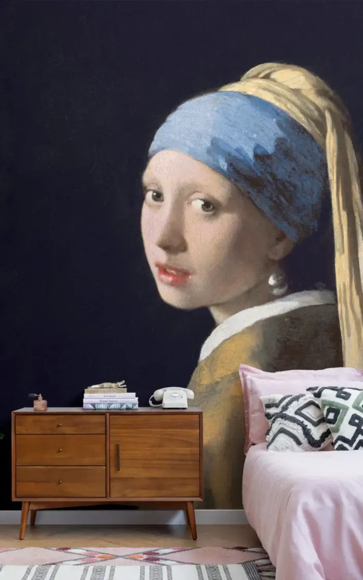 A bedroom featuring a large mural of "Girl with a Pearl Earring" on the wall above a mid-century modern wooden dresser, beside a bed with pink bedding and a patterned throw pillow.