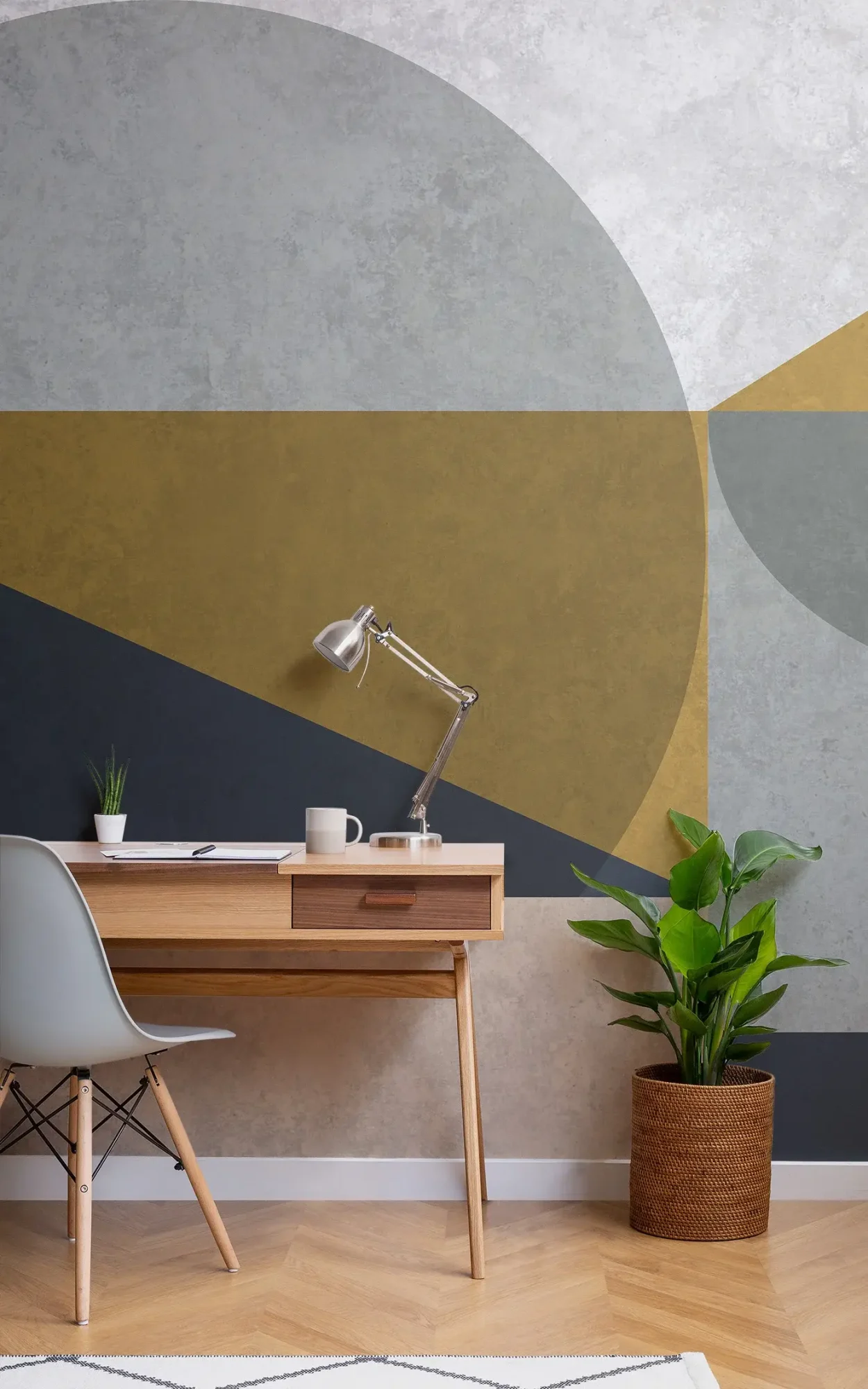 A modern home office setup featuring a minimalist desk with a wooden top and white chair, a silver desk lamp, and a large geometric patterned wallpaper in shades of gold, grey, and black, accompanied by a potted plant in a woven basket.