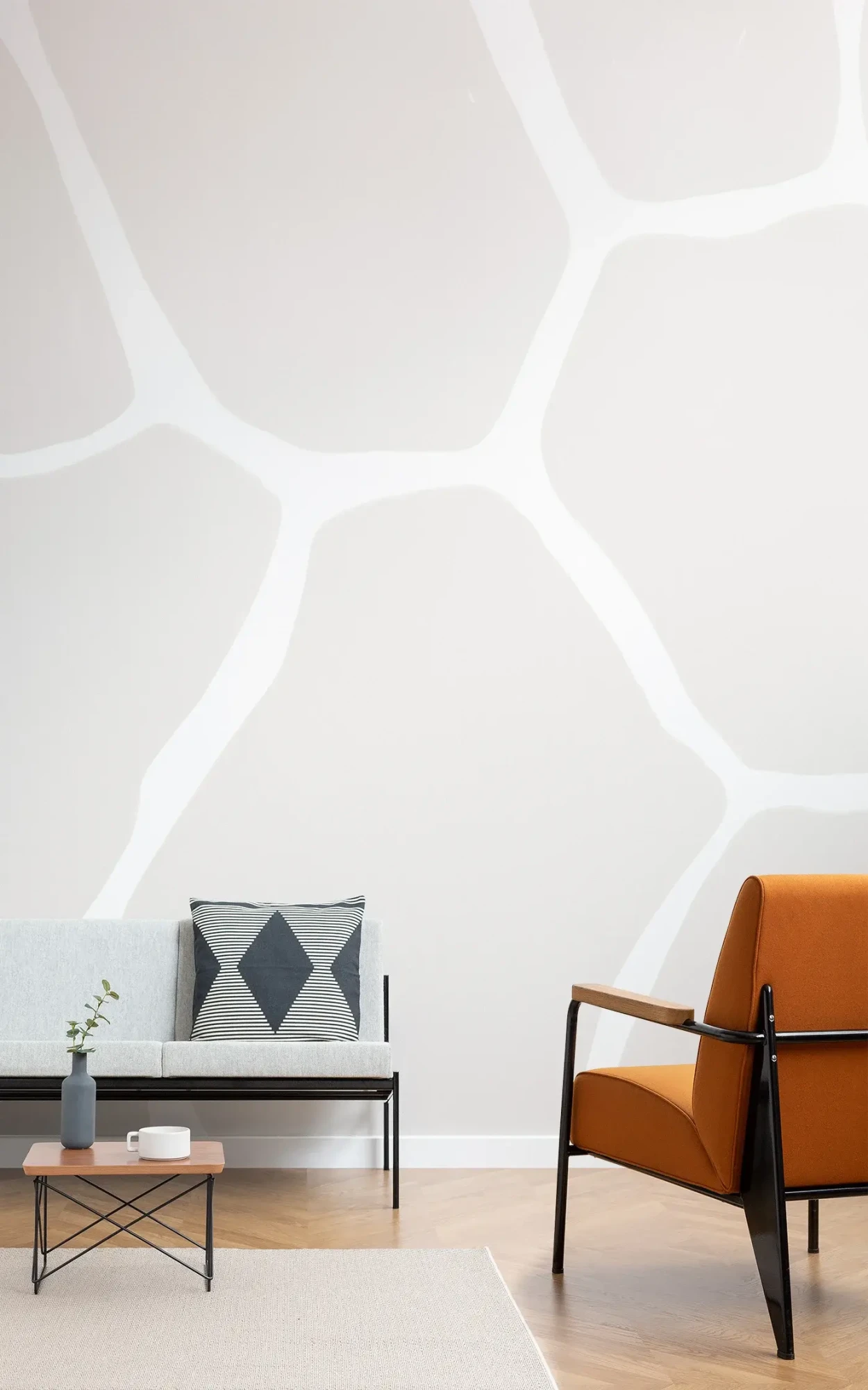 A modern living space with a large white wall featuring an abstract, organic-shaped grey pattern, furnished with a grey sofa with a geometric pillow, a burnt orange armchair, a small wood and metal coffee table, and a beige rug.
