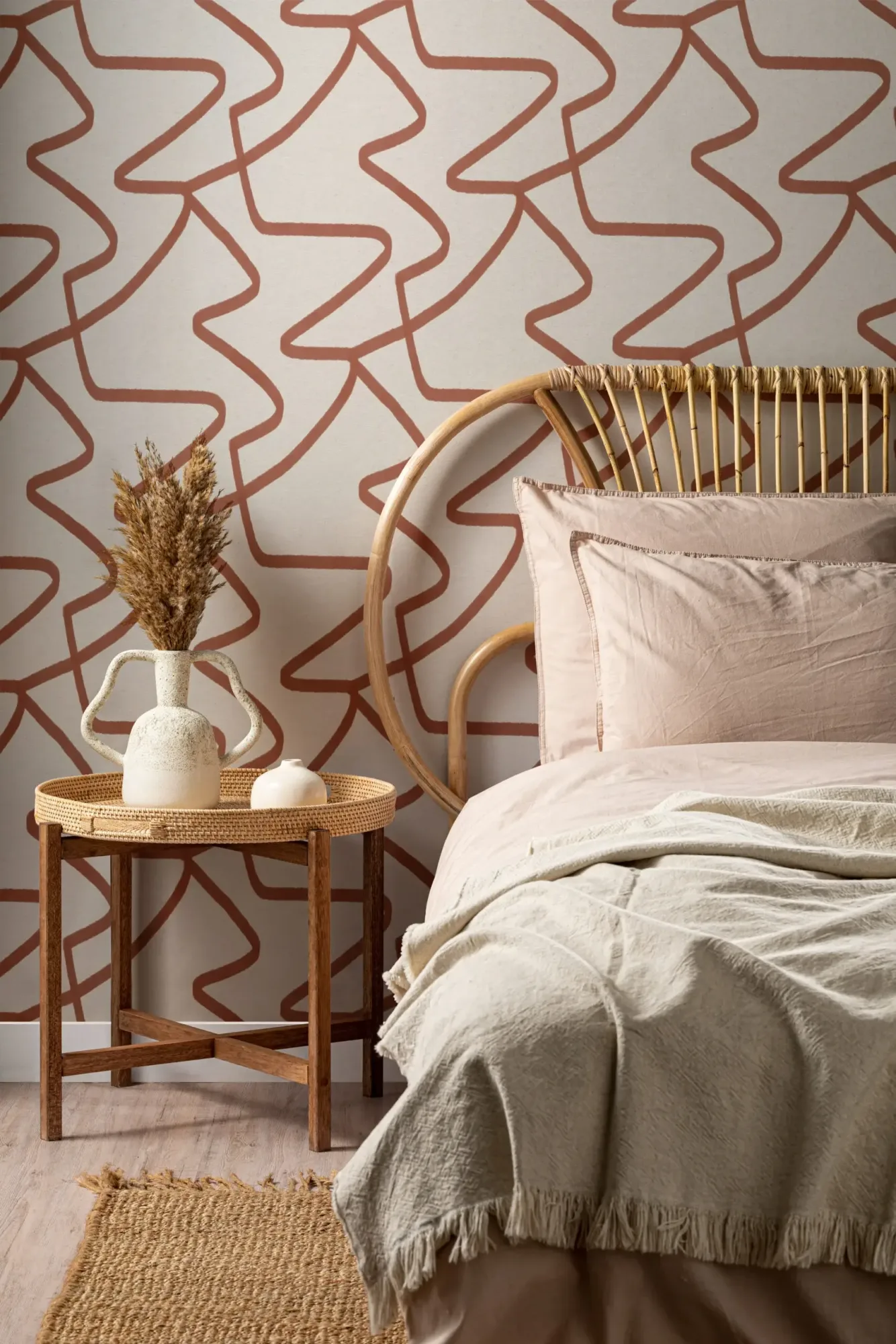 A cozy bedroom setting featuring a natural rattan bed with soft pink bedding, a rattan side table with a rustic vase and dried flowers, against a cream wallpaper adorned with a terracotta abstract line pattern.