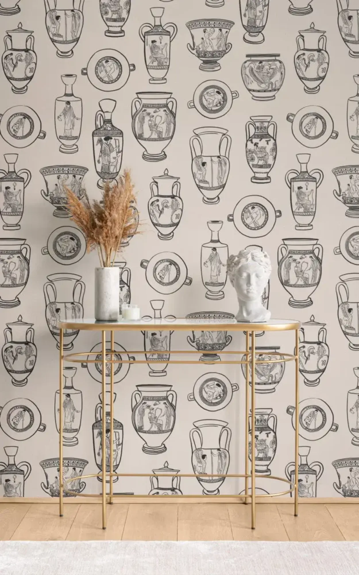  An elegant room decor featuring a wallpaper with classical Greek vase illustrations, complemented by a gold console table adorned with a vase of dried pampas grass, a classical bust sculpture, and decorative ceramics.