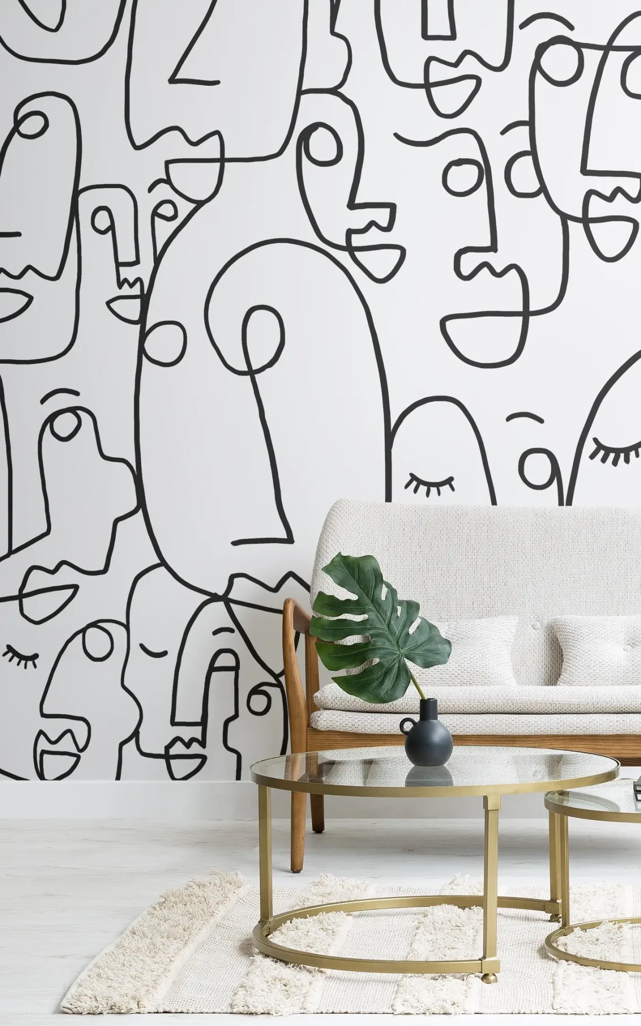A contemporary living space featuring a beige sofa with a large monstera leaf on a coffee table, against a bold black and white abstract face pattern wallpaper.