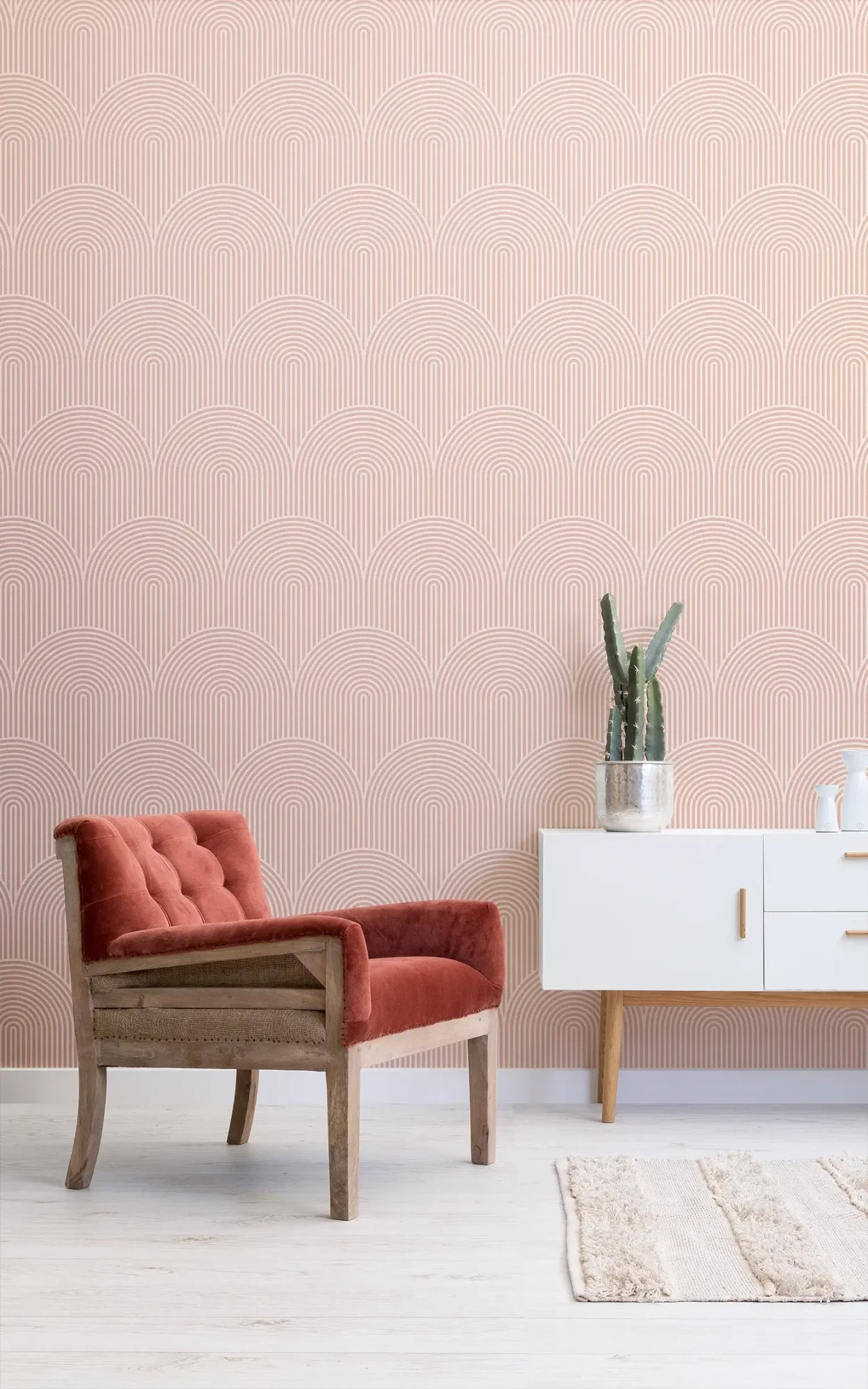  A modern interior design featuring a red velvet armchair against a subtle pink wallpaper with an art deco style pattern, accompanied by a white sideboard and a cactus plant.