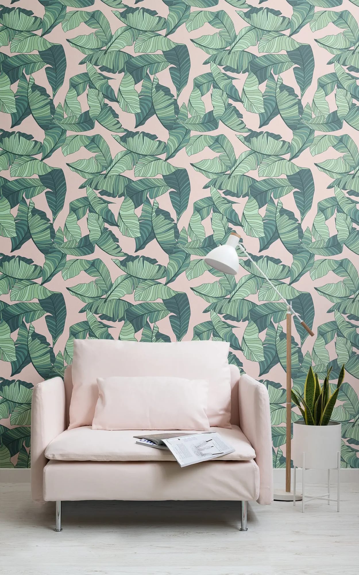 A minimalist living room with a soft pink sofa, a floor lamp, and a pot with a green plant, set against a pastel pink wallpaper adorned with large green banana leaves.