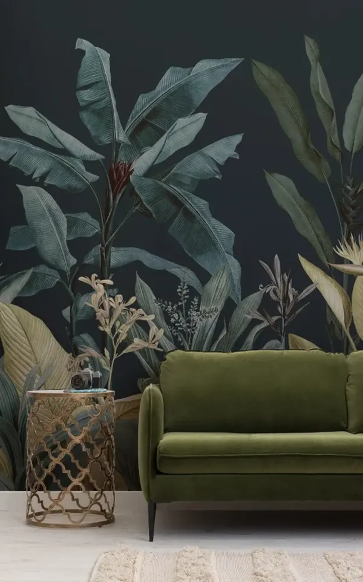 An elegant living room featuring a plush green velvet sofa, a decorative gold side table, and a large mural of dark tropical plants on a navy blue background.