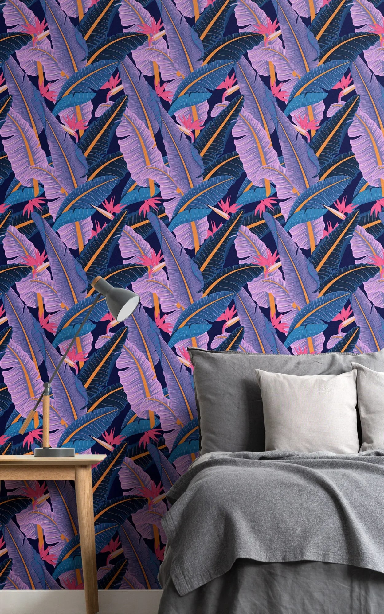 A modern bedroom featuring a gray bed with matching linens against a vibrant tropical-themed wallpaper with pink and purple leaves on a dark blue background.