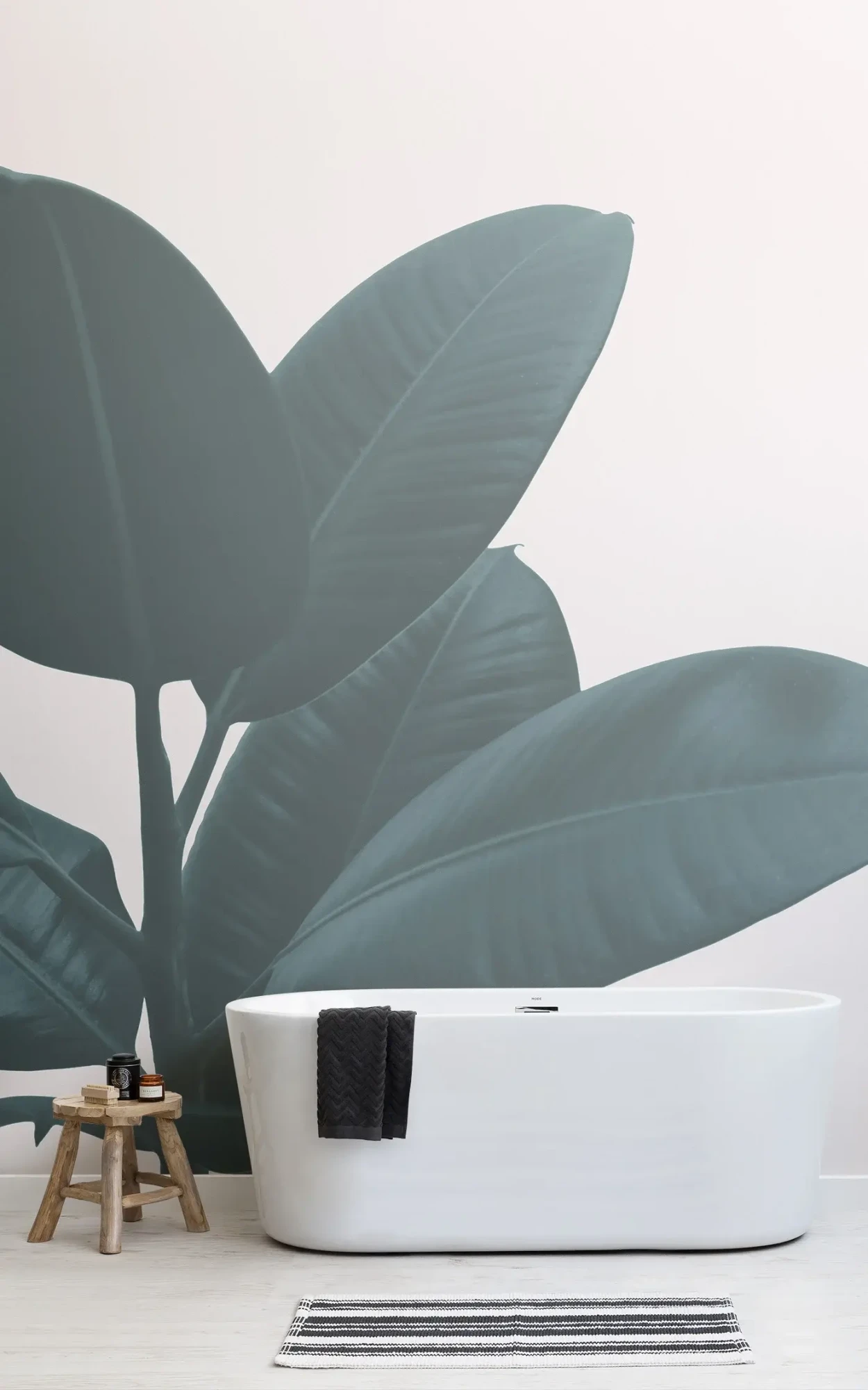 A modern bathroom features a freestanding white bathtub, a small wooden stool with bath products, and a large, dark green leaf mural on the wall behind.