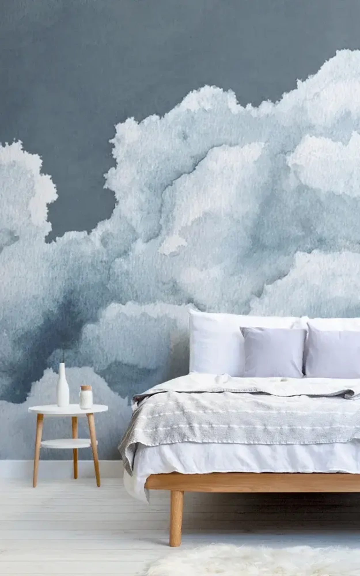 A serene bedroom features a wooden bed with white and gray bedding, a small side table with decor items, and a mural of a dark blue cloudy sky on the wall behind.
