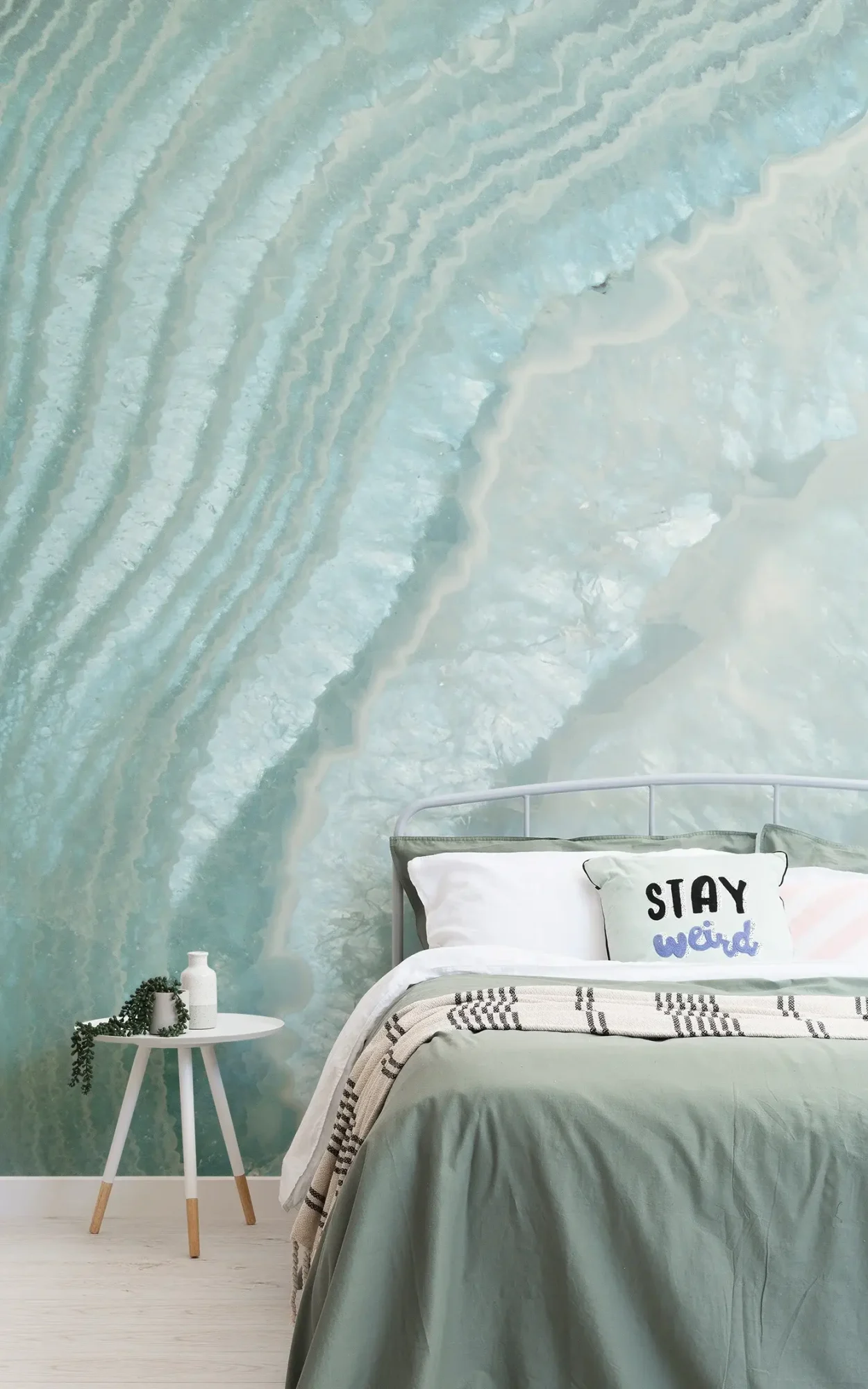  A stylish bedroom features a metal bed with white and green bedding, a "Stay Weird" pillow, a small side table with decor, and a mural of light blue agate on the wall behind.