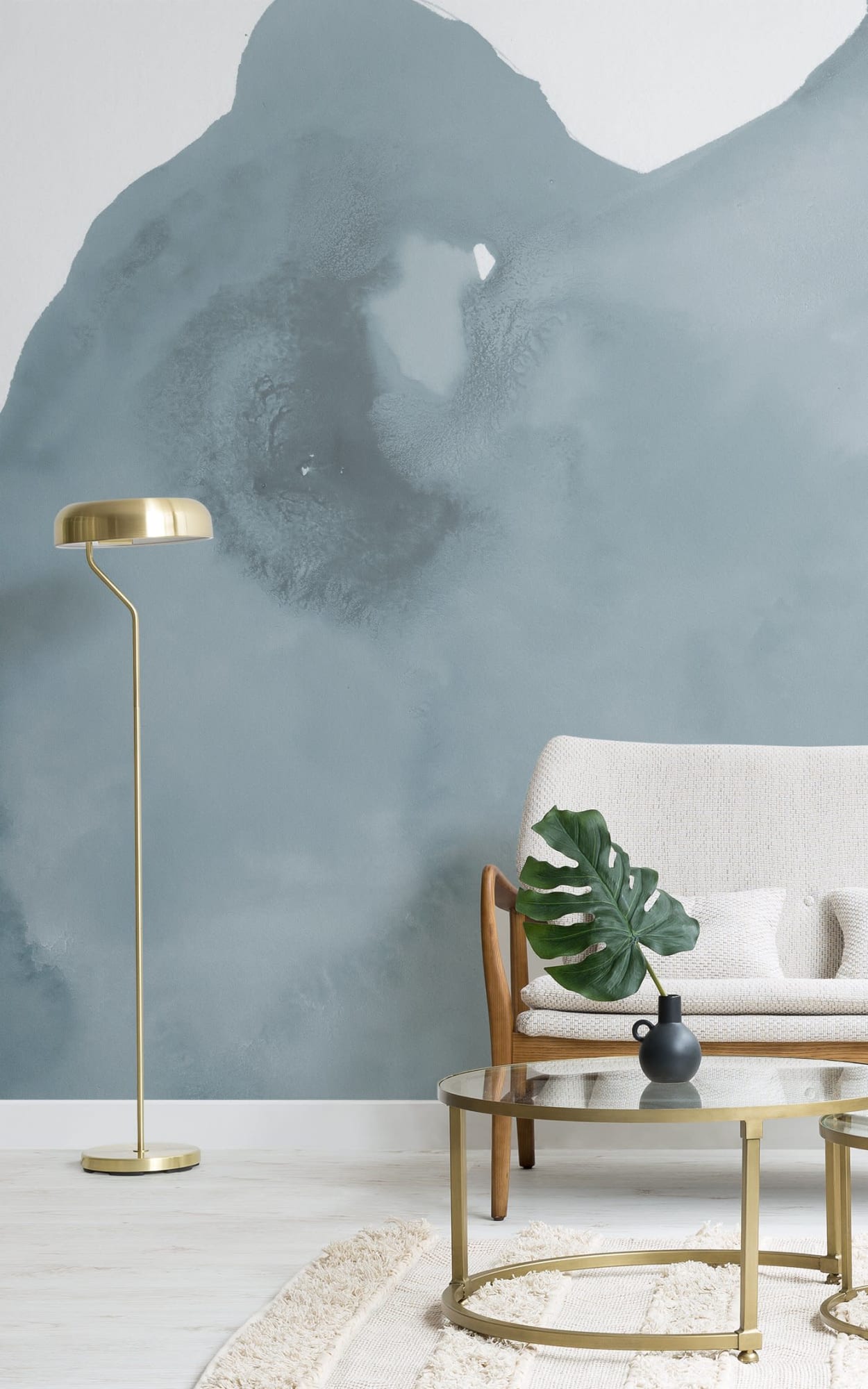  A stylish living room features a beige armchair, a glass coffee table with a black vase holding a green leaf, a brass floor lamp, and a blue abstract mural on the wall behind.