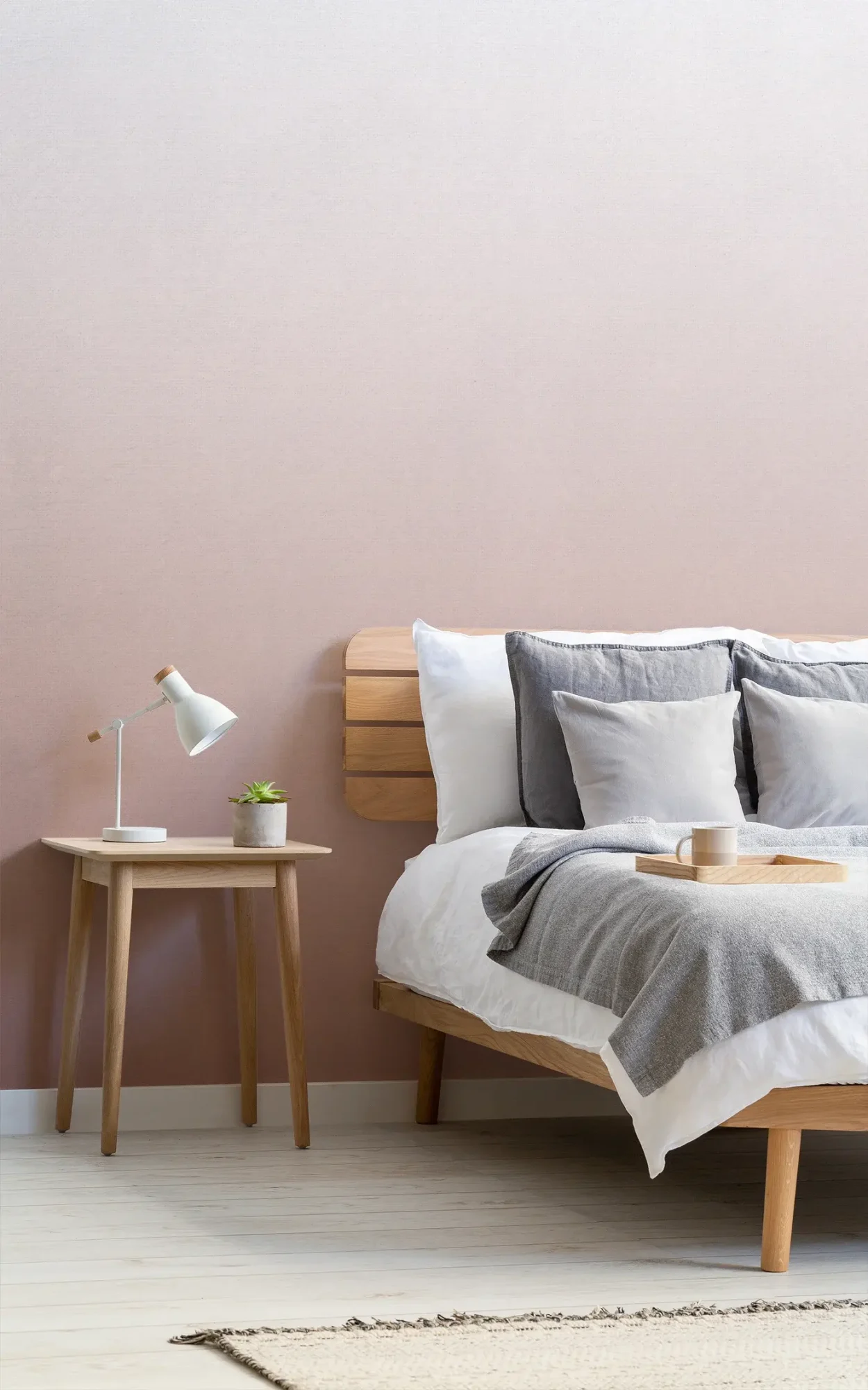  A minimalist bedroom features a wooden bed with white and gray bedding, a wooden side table with a lamp and plant, and a soft pink ombre wall behind.