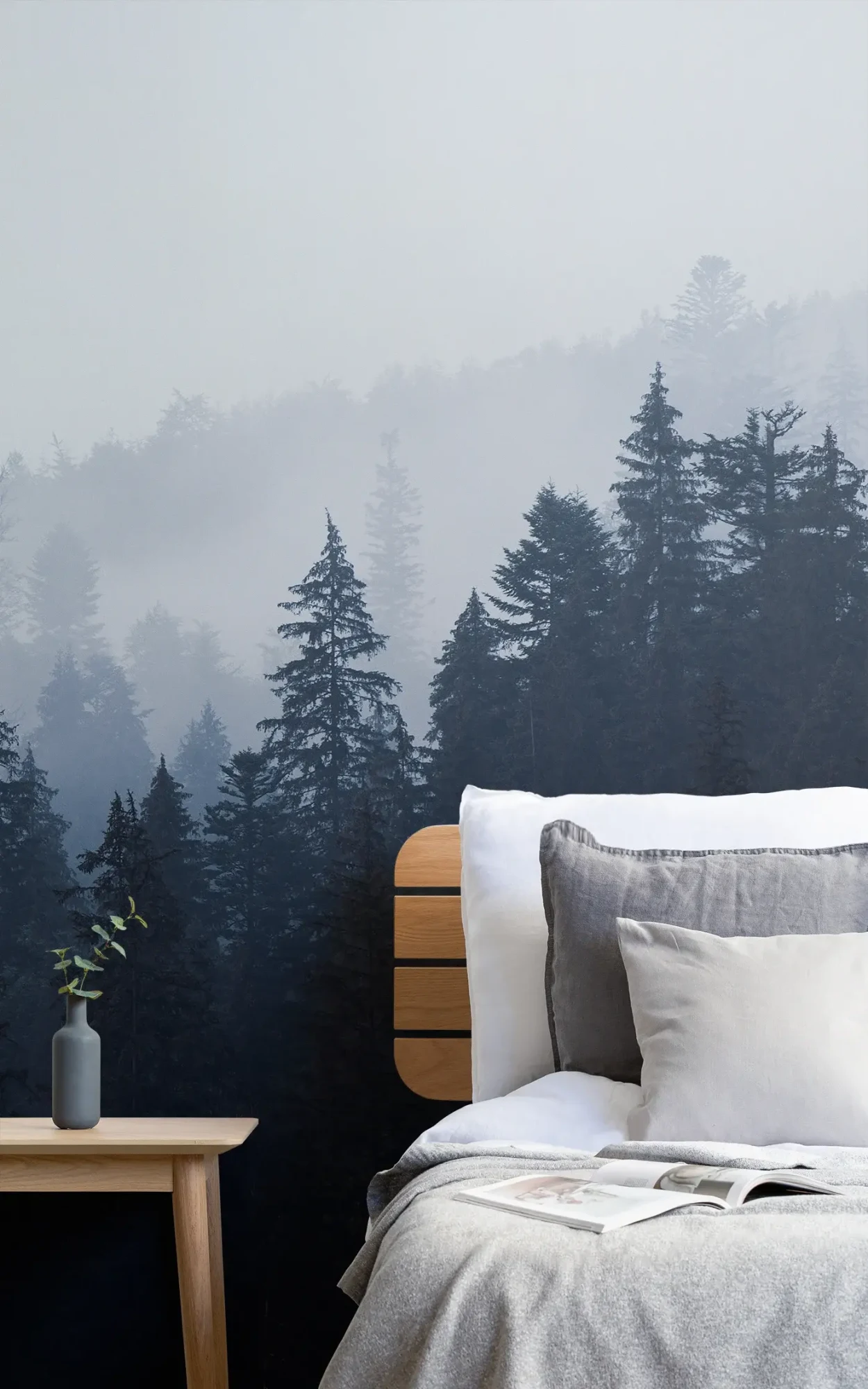  A cozy bedroom features a wooden bed with white and gray bedding, a small wooden table with a vase, and a mural of misty pine trees on the wall behind.