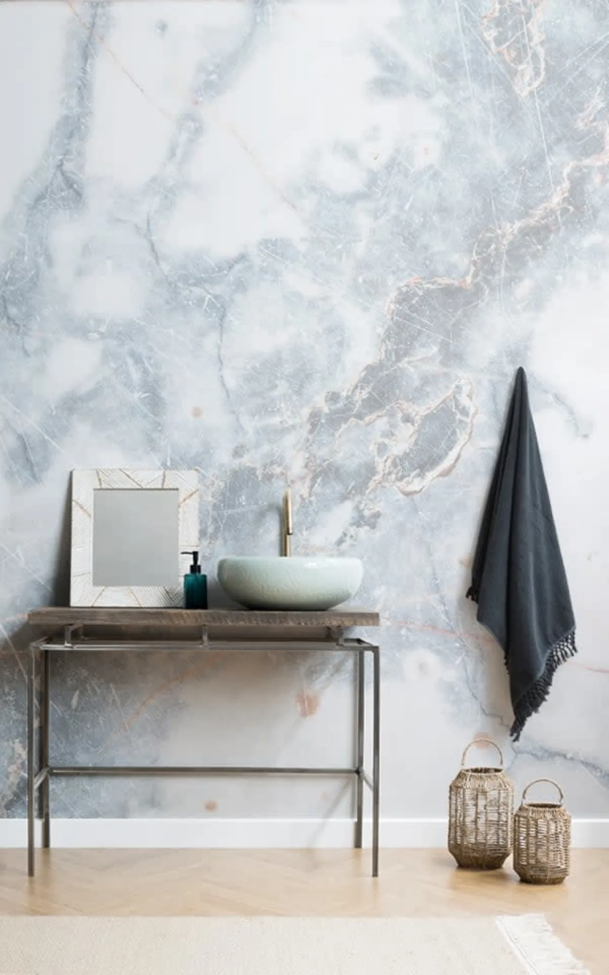 A modern bathroom features a wooden and metal vanity with a green vessel sink, a framed mirror, a hanging towel, and a light blue marble wall mural behind.