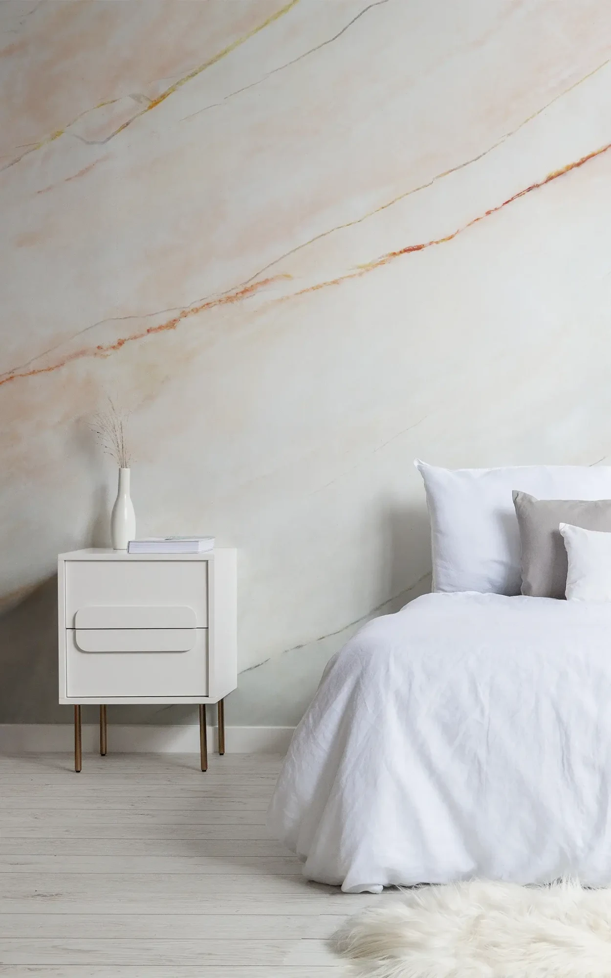  A minimalist bedroom features a white bed, a white nightstand with a vase and books, and a marble-textured wall mural behind.