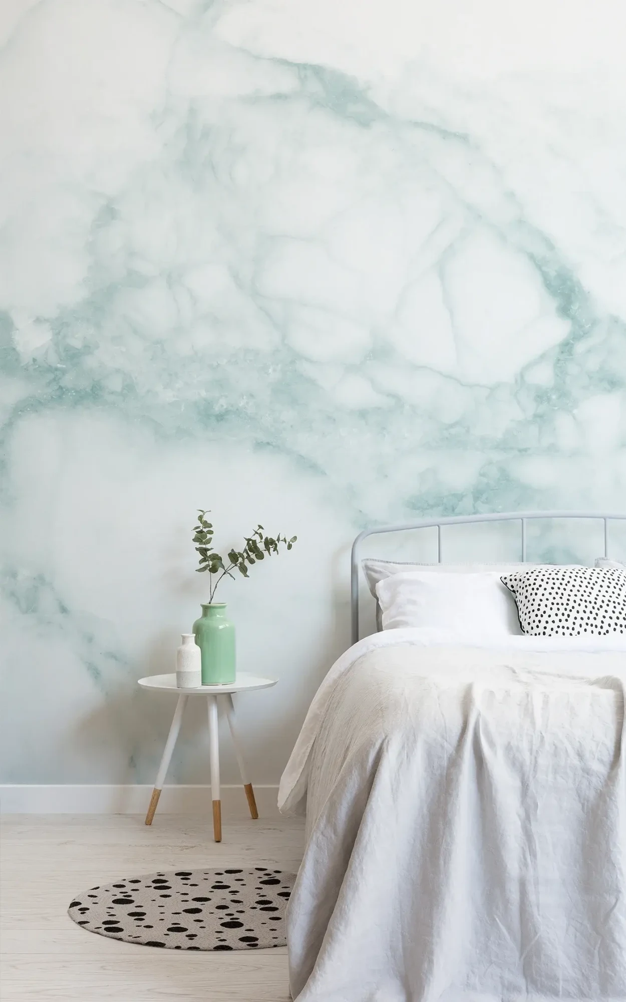 A minimalist bedroom features a white bed, a side table with green and white vases, and a wall mural with a green marble pattern behind.
