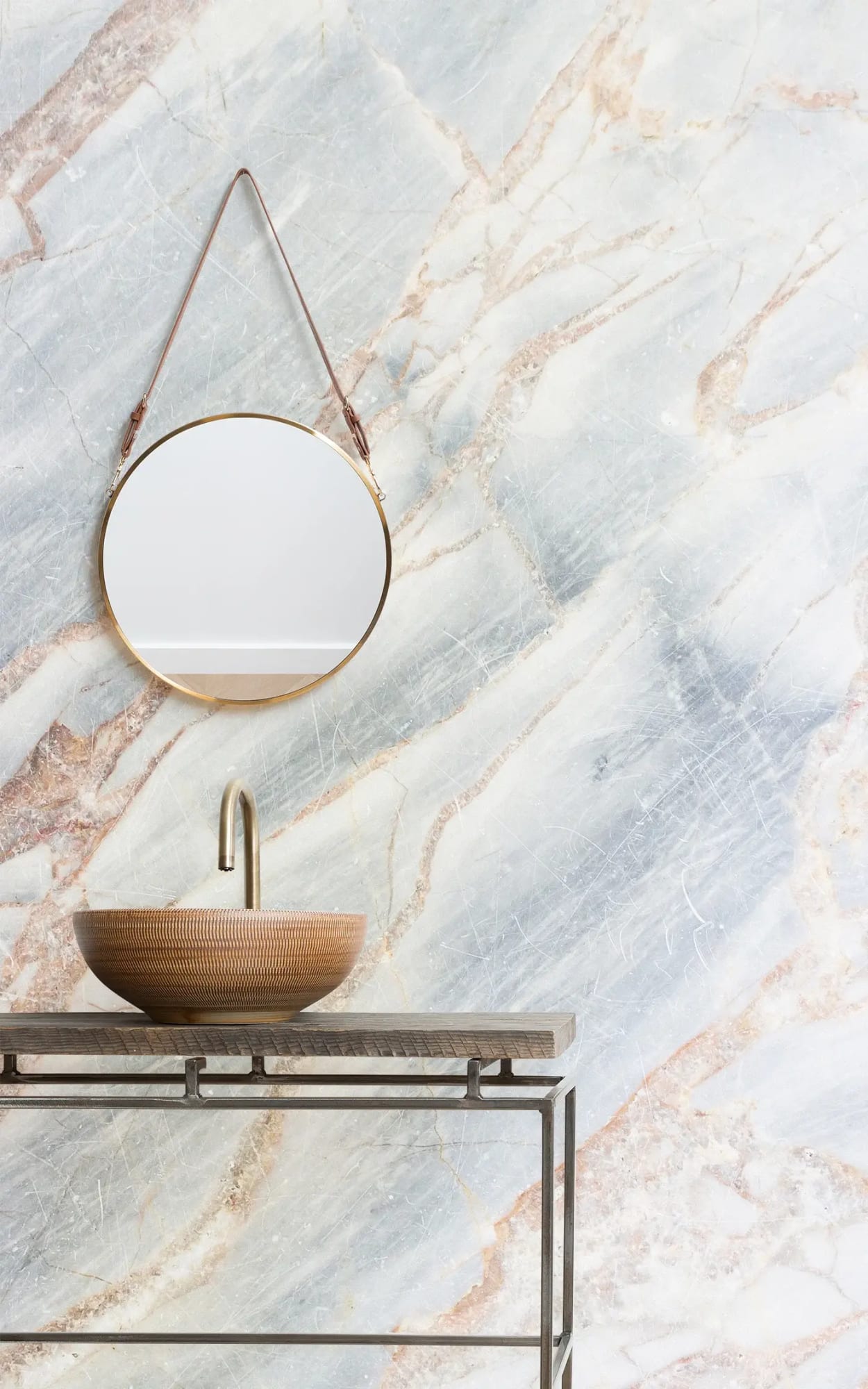 A modern bathroom features a round mirror with a leather strap, a stone vessel sink on a metal and wood vanity, and a marble wall mural behind.
