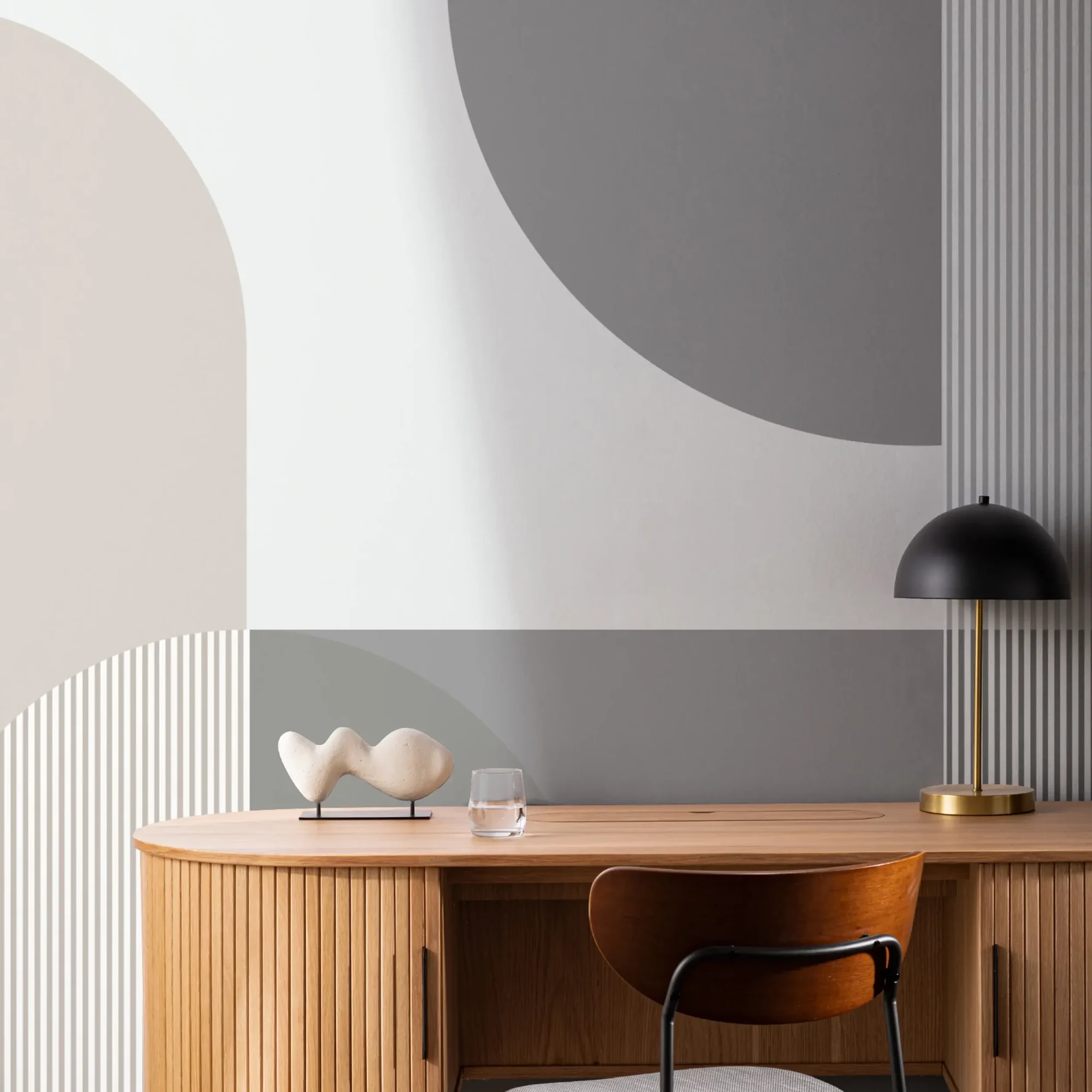 A modern office features a wooden desk with a decorative sculpture, a glass of water, a black lamp, a wooden chair, and a geometric wallpaper mural in shades of gray and beige behind.