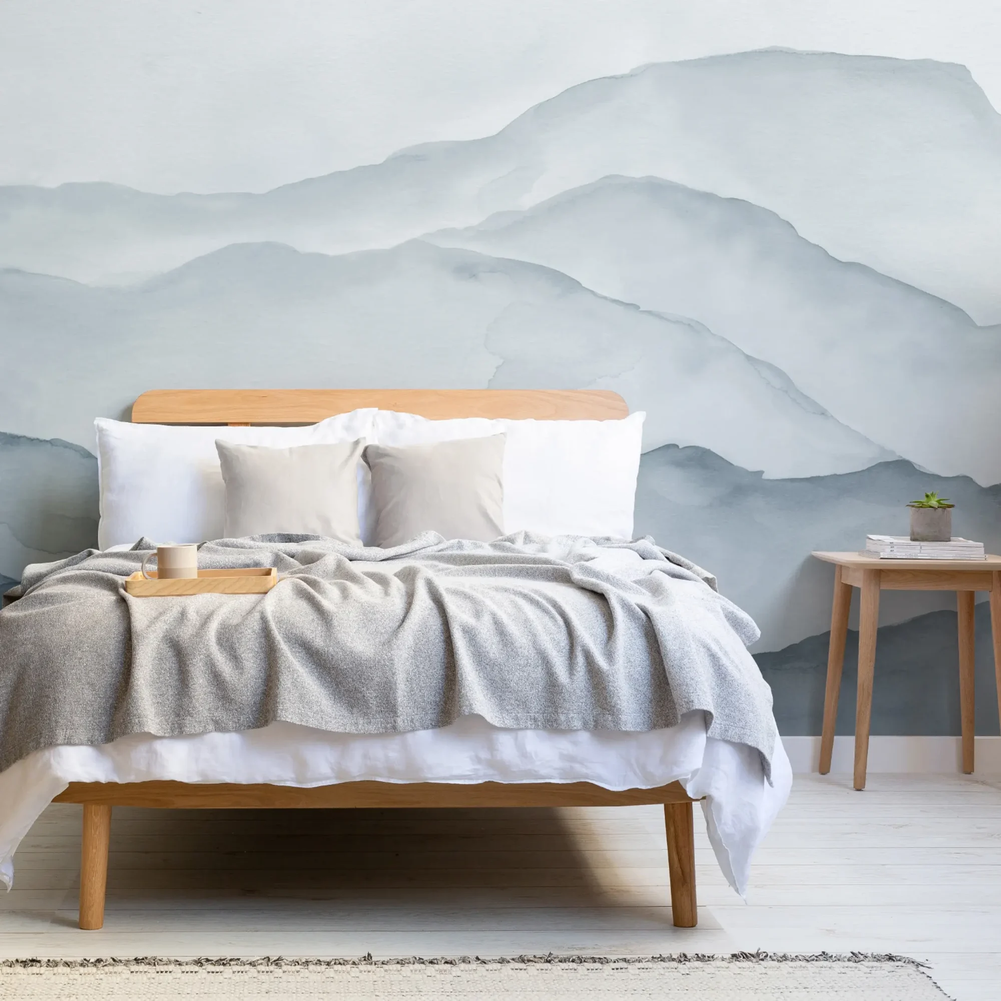A minimalist bedroom features a wooden bed with white and gray bedding, a wooden side table, and a mural of blue watercolor mountains on the wall behind.