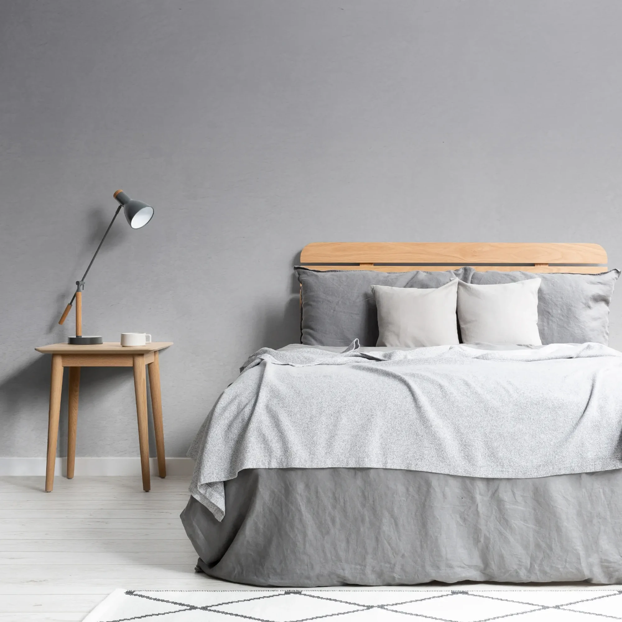  A minimalist bedroom features a wooden bed with gray and white bedding, a wooden side table with a lamp and mug, and a gray ombre wall behind.