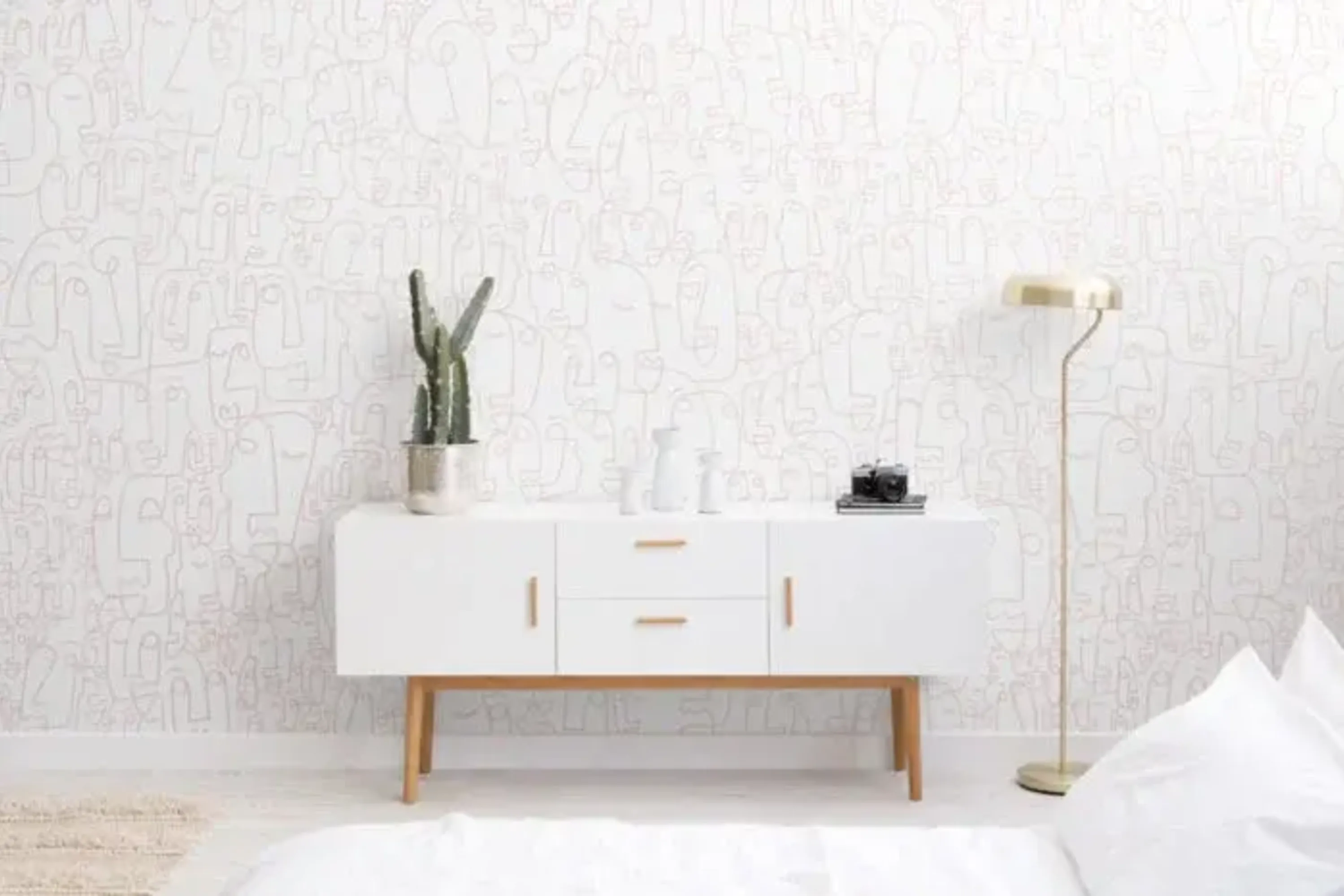  A minimalist room features a white sideboard with decor items, a gold floor lamp, a potted cactus, and a white wall with pink face doodles.