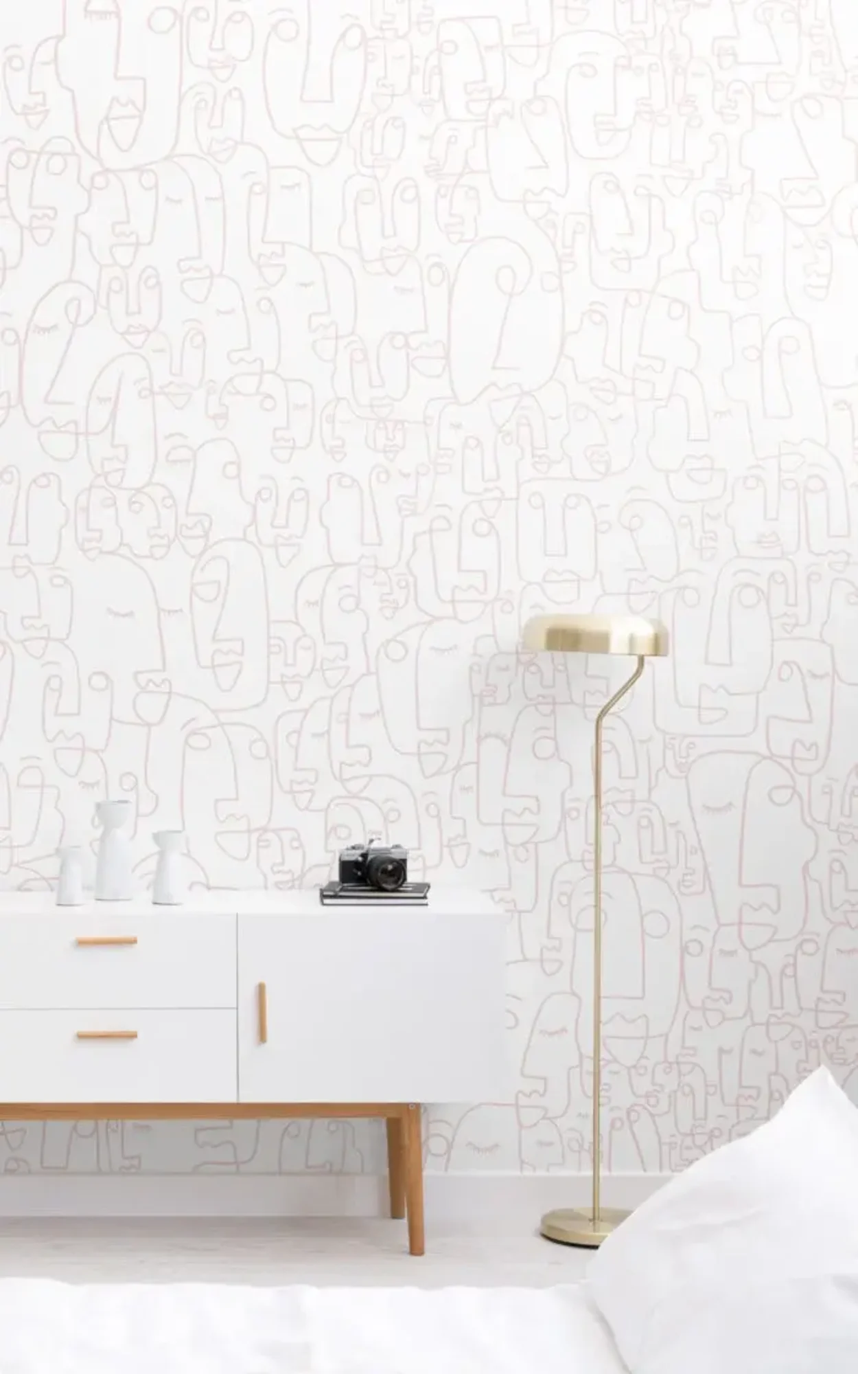  A modern room features a white sideboard with decorative items, a gold floor lamp, and a white wall with a pink face doodle pattern.