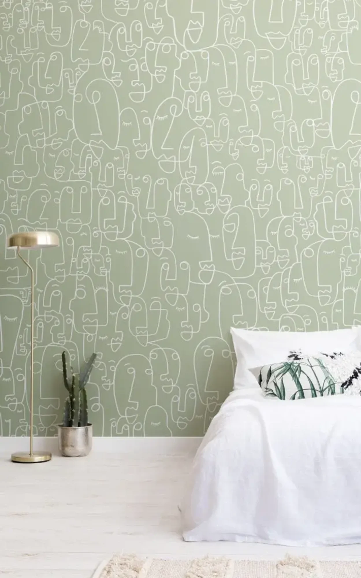  A minimalist room features a white bed with decorative pillows, a gold floor lamp, a potted cactus, and a green wall with white face doodles.
