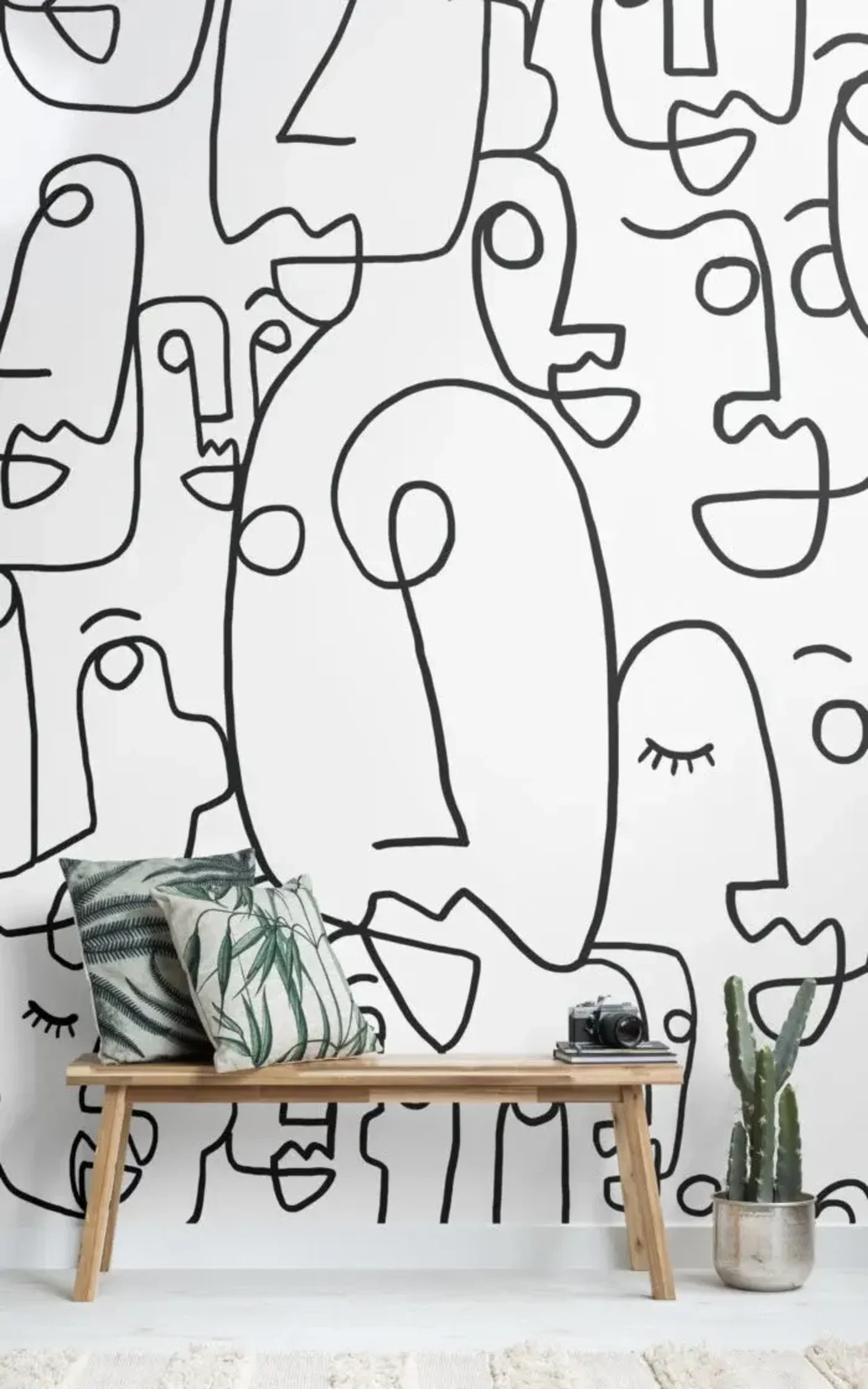 A modern room features a wooden bench with decorative pillows, a potted cactus, and a white wall with large black face doodles.