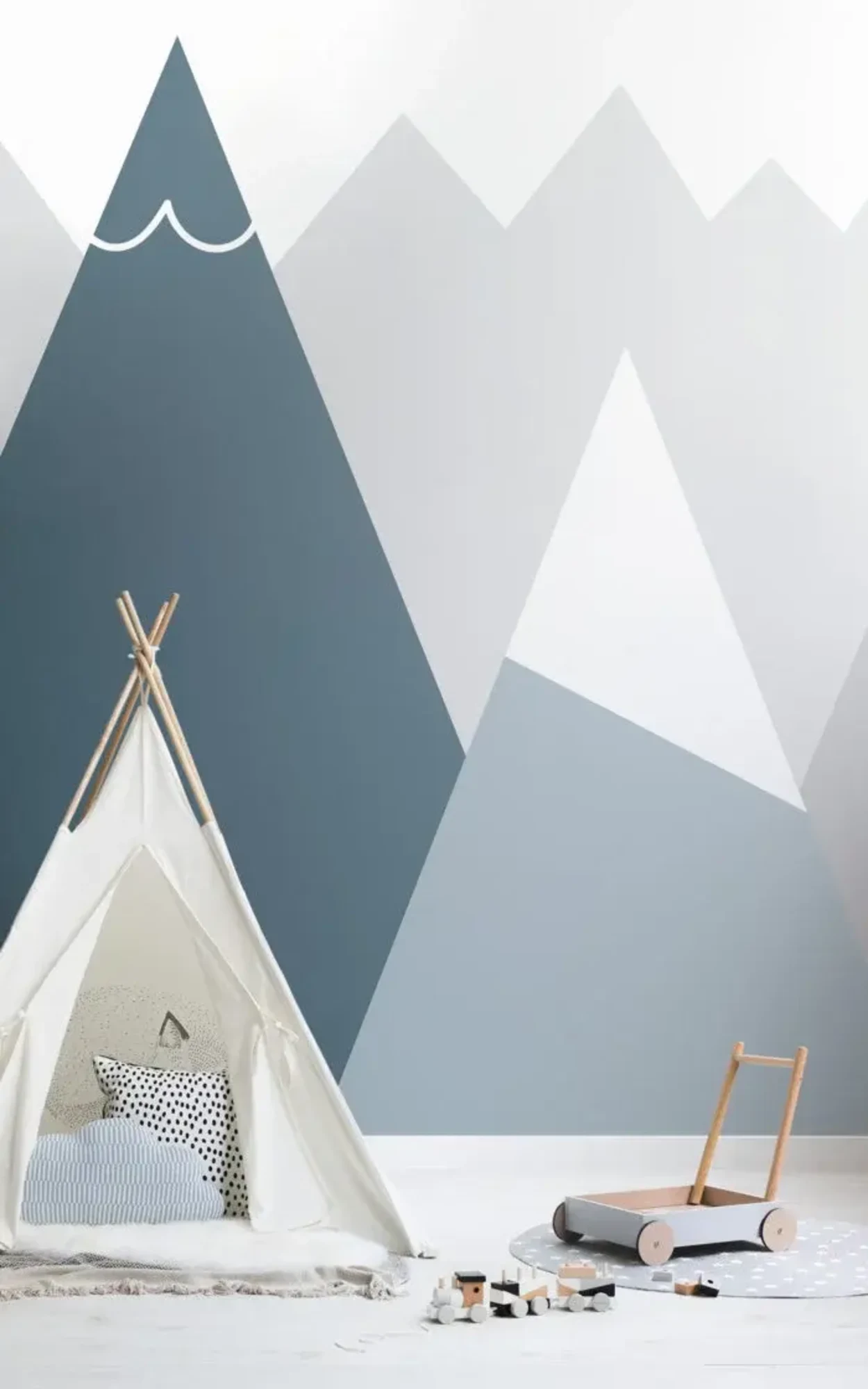 A children's room features a play tent with pillows, a toy train on the floor, and a mural of geometric mountains in shades of blue and gray on the wall behind.