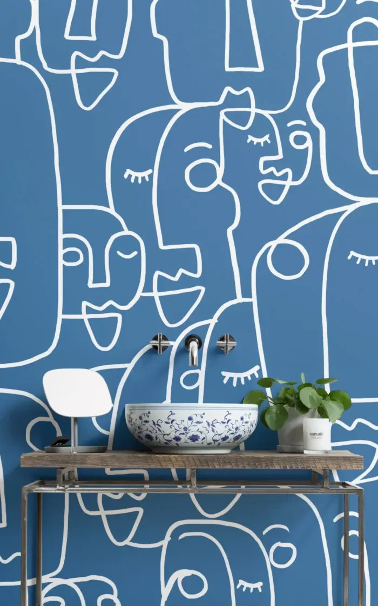 A stylish bathroom features a blue wall with white abstract face outlines, a decorative blue and white sink, a modern faucet, and a small plant on a metal console table.