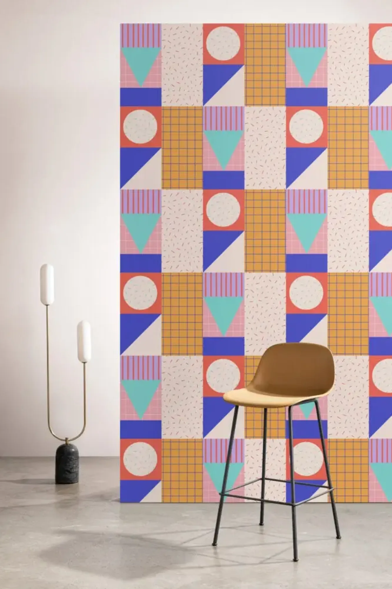 A modern interior features a colorful geometric patterned wall, a minimalist floor lamp with two vertical lights, and a brown barstool with a black metal frame.