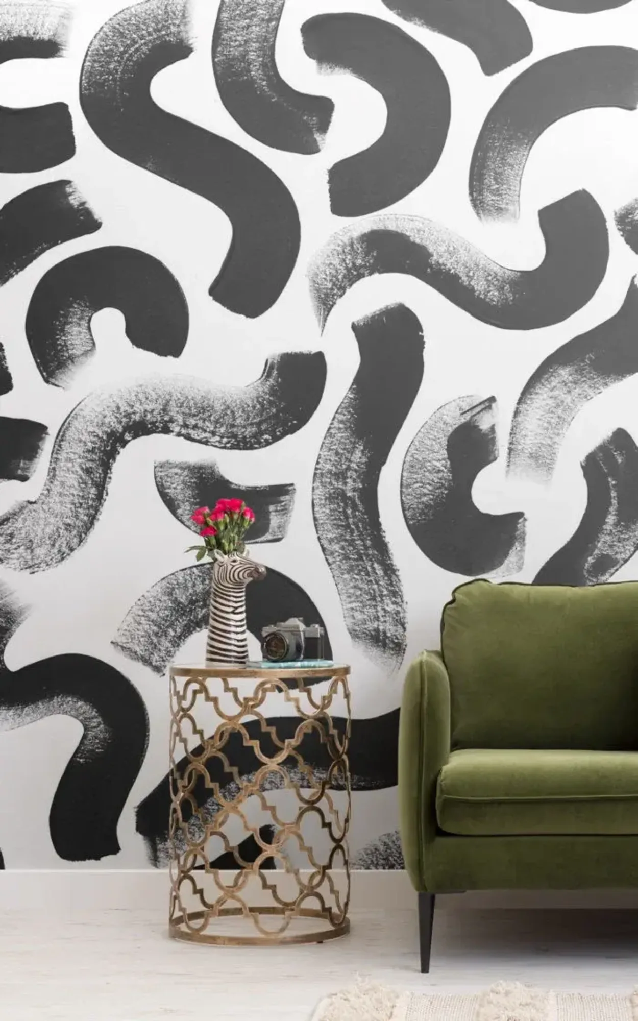 A modern living room features a green sofa, an ornate side table with a zebra-shaped vase and pink flowers, and a black-and-white abstract patterned wall.