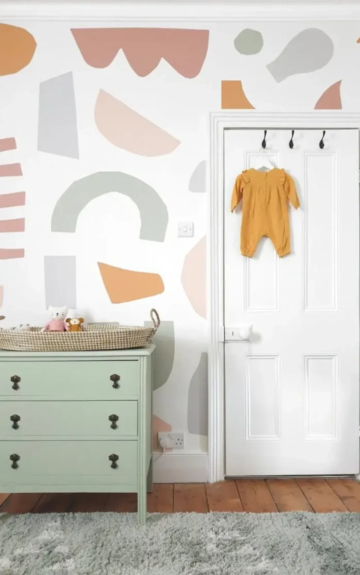  A nursery features a mint green dresser with a changing basket, a colorful abstract patterned wall, and a white door with a yellow baby outfit hanging on it.