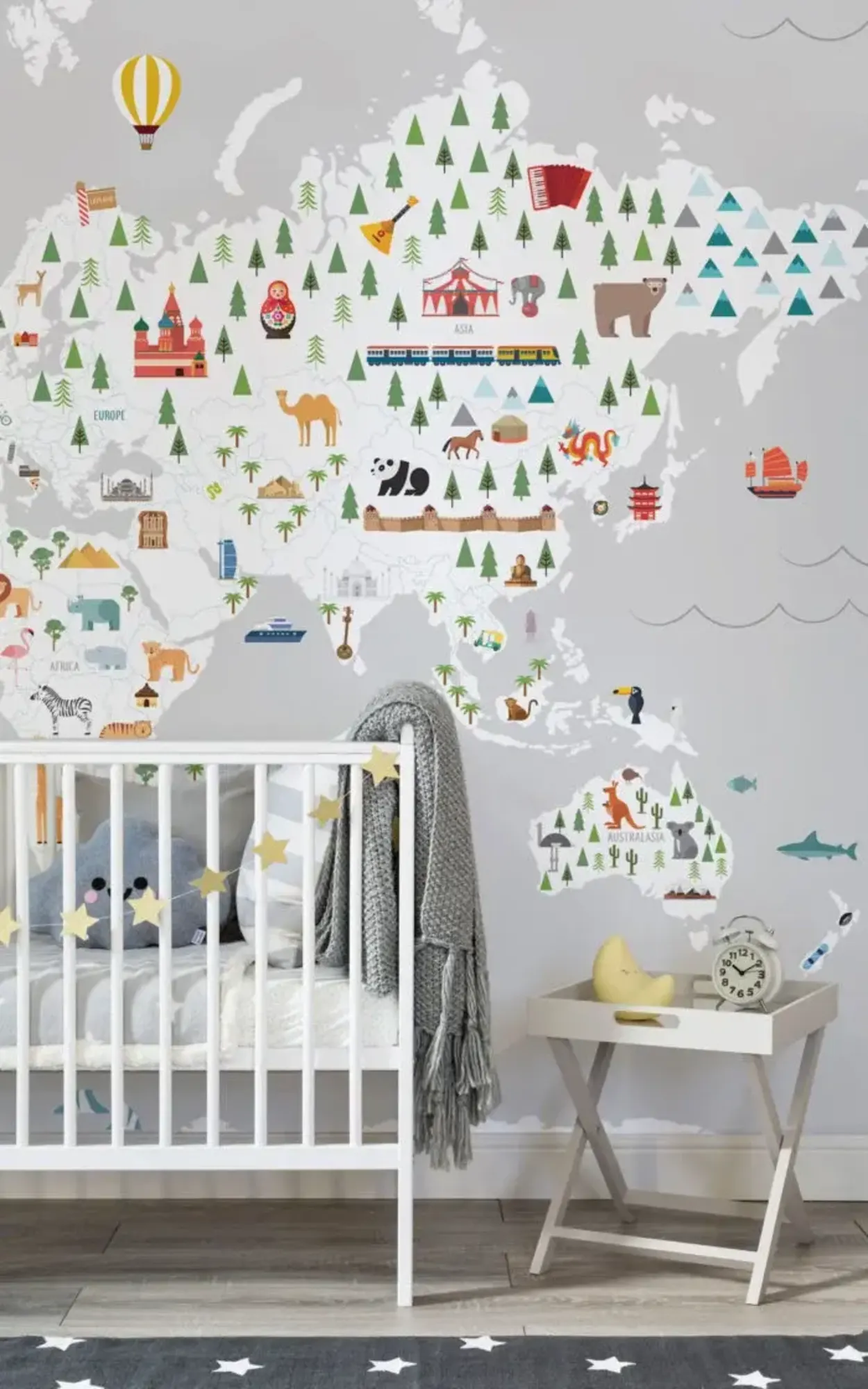  A nursery features a white crib with a stuffed pillow and blanket, a side table with a clock and decorative items, and a colorful world map mural on the wall.