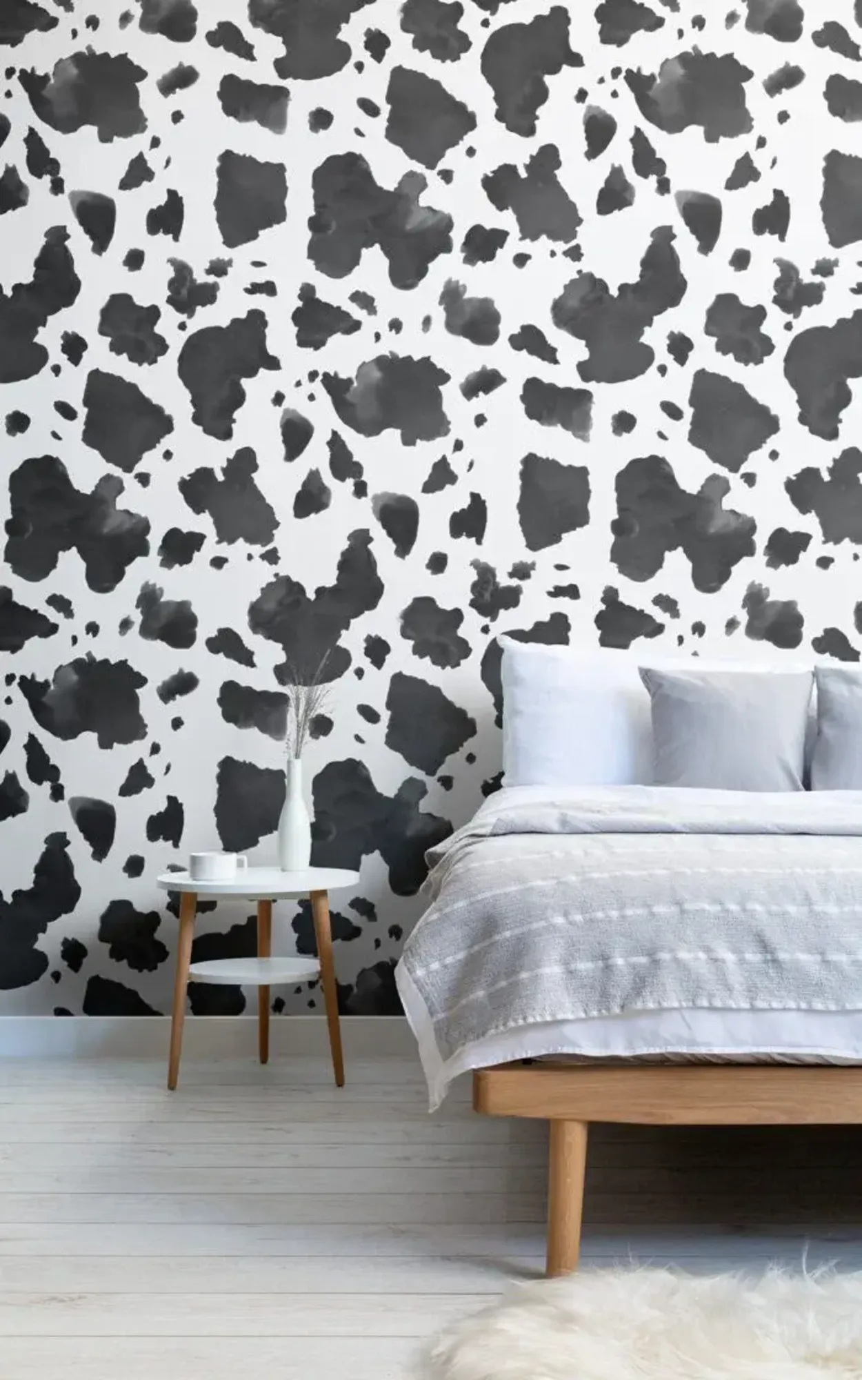  A minimalist bedroom features a black and white cow print wallpaper, a wooden bed with gray and white bedding, and a small round side table with decor.