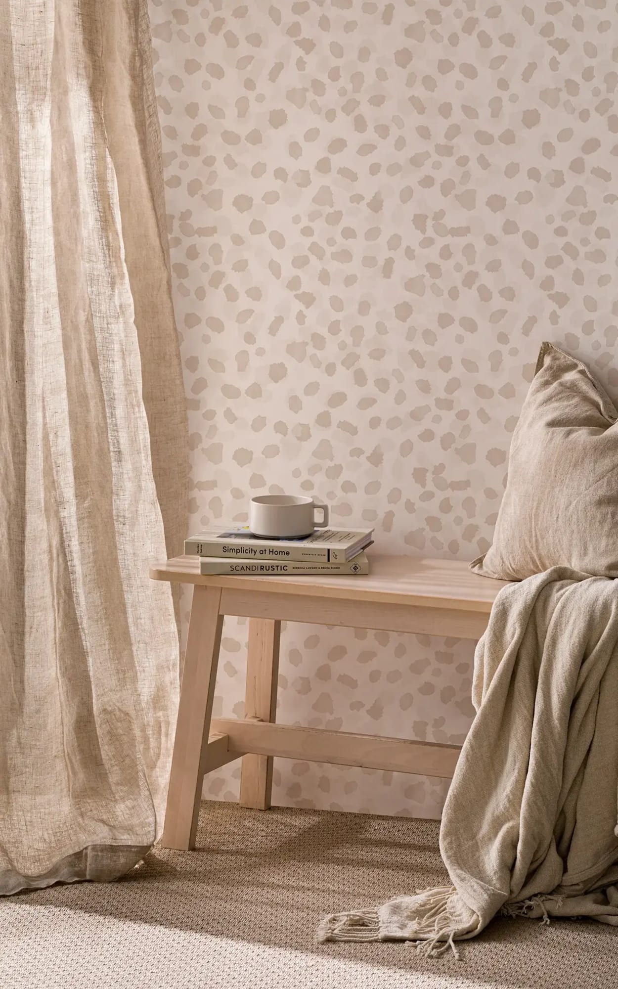 A cozy reading nook features a light wooden bench with books and a cup, beige pillows, a blanket, and a wall with subtle beige cheetah print wallpaper.