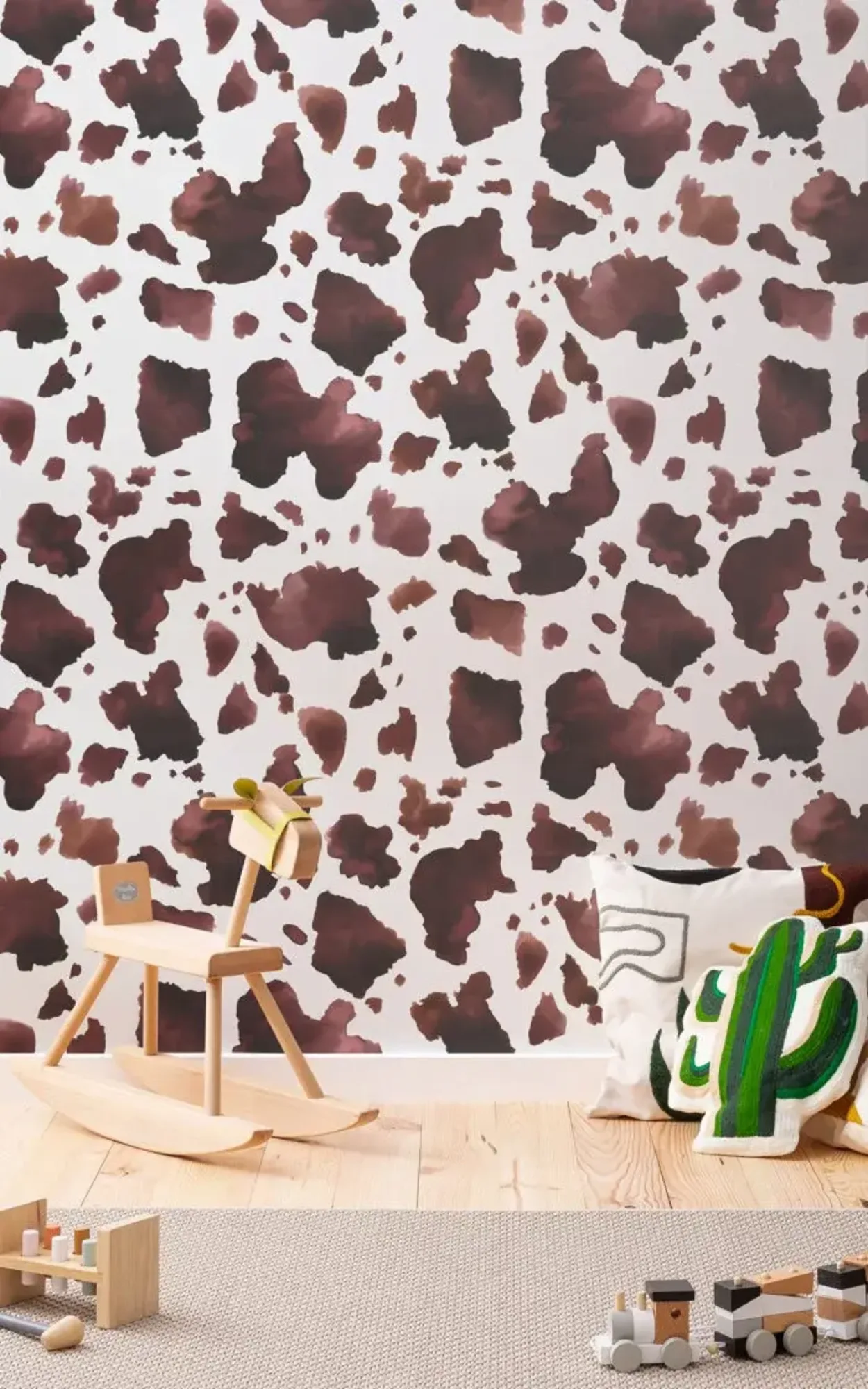 A children's room features a wooden rocking horse, decorative pillows, and toys, set against a wall with a brown cow print pattern.