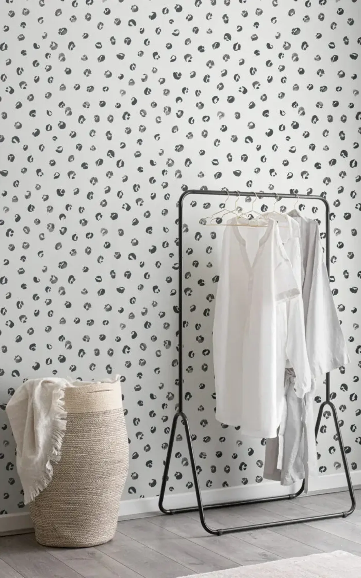 A minimalist room features a clothing rack with white garments, a woven basket with a blanket, and a wall with a black and white leopard print pattern.