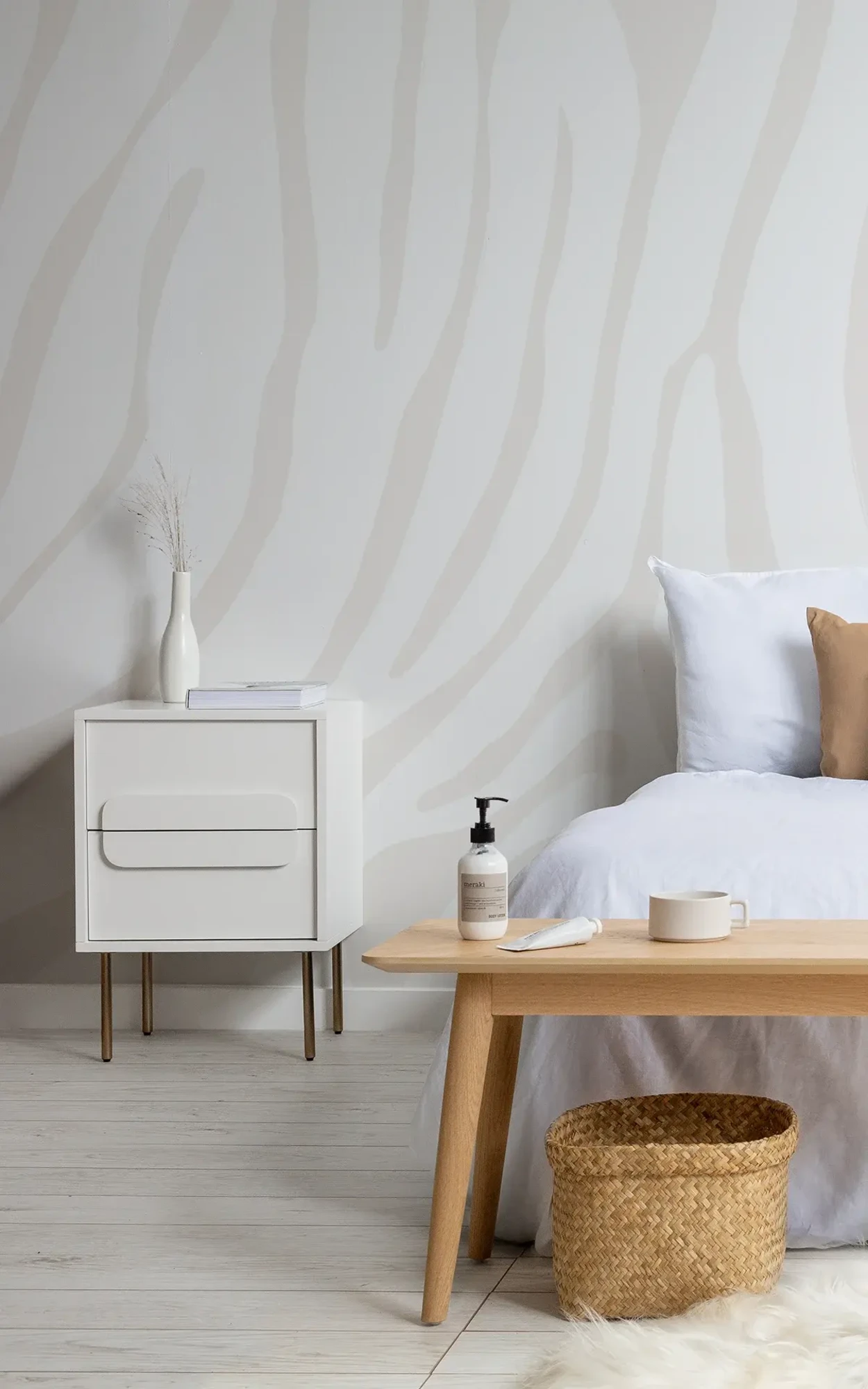 A minimalist bedroom features a white nightstand, a wooden bench, and a wall with light beige zebra stripes, complemented by white bedding and neutral decor.