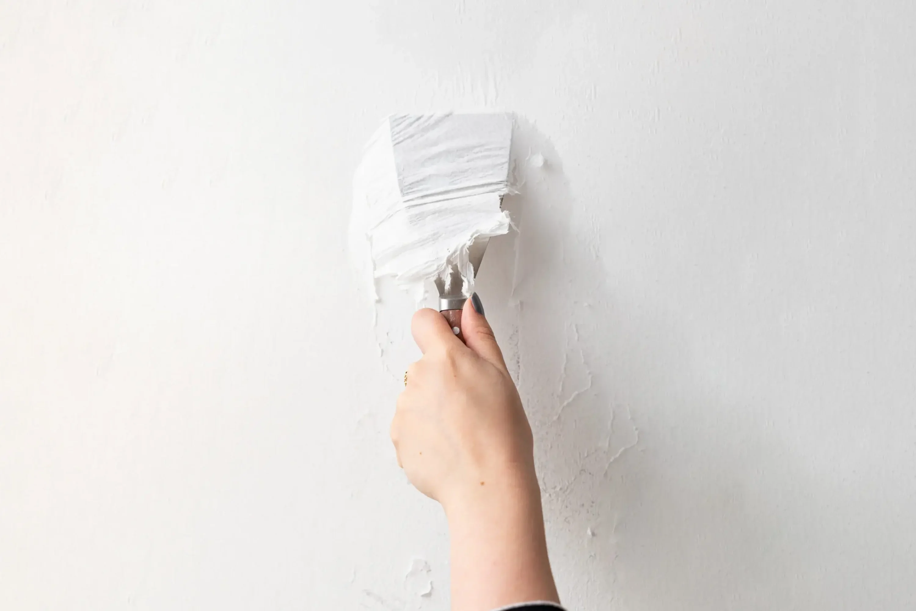 A person scrapes off wallpaper glue from a wall using a putty knife.