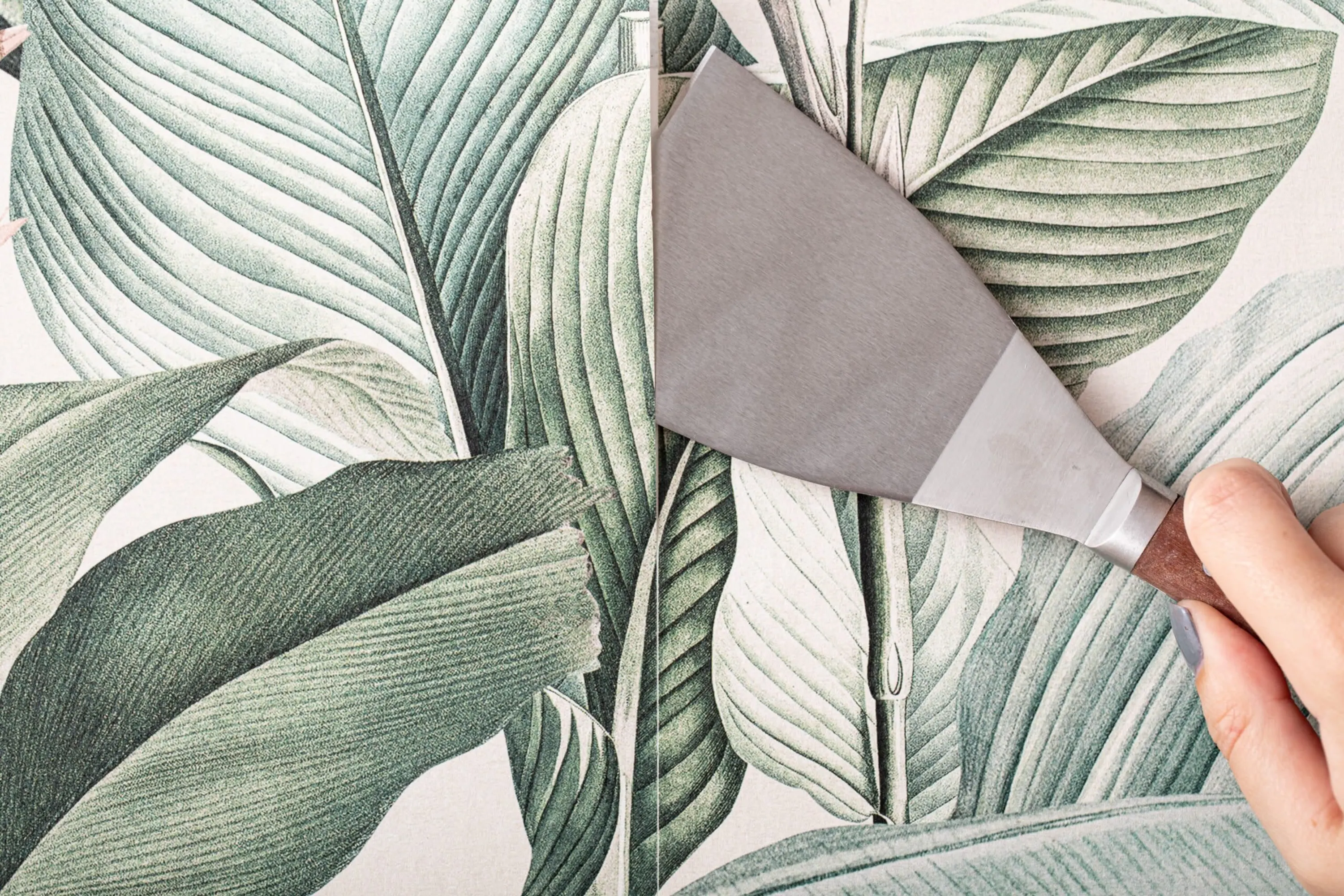 A hand using a putty knife to peel off wallpaper with a green leaf pattern.