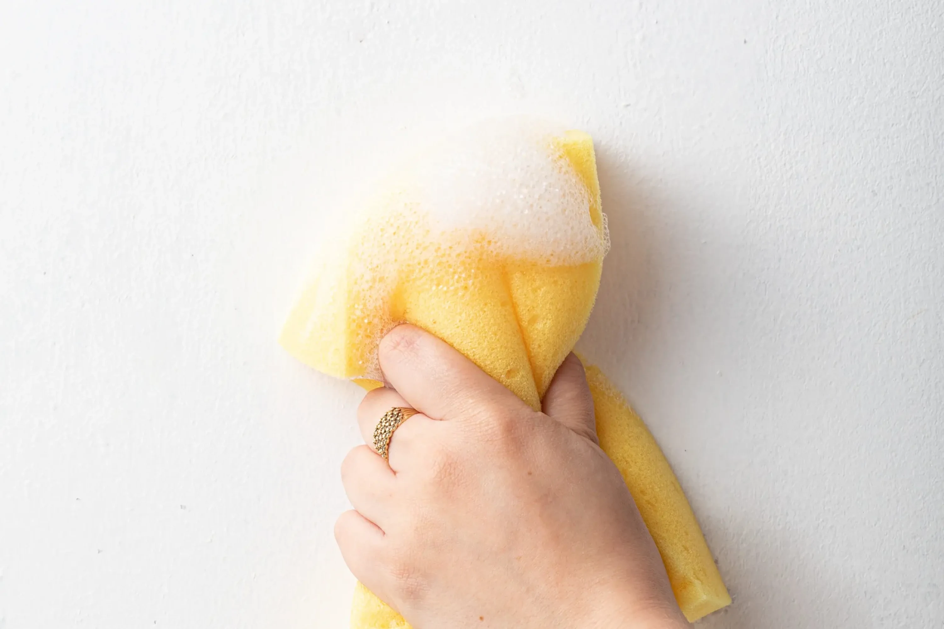 A person cleans a white wall with a yellow sponge and soapy water.