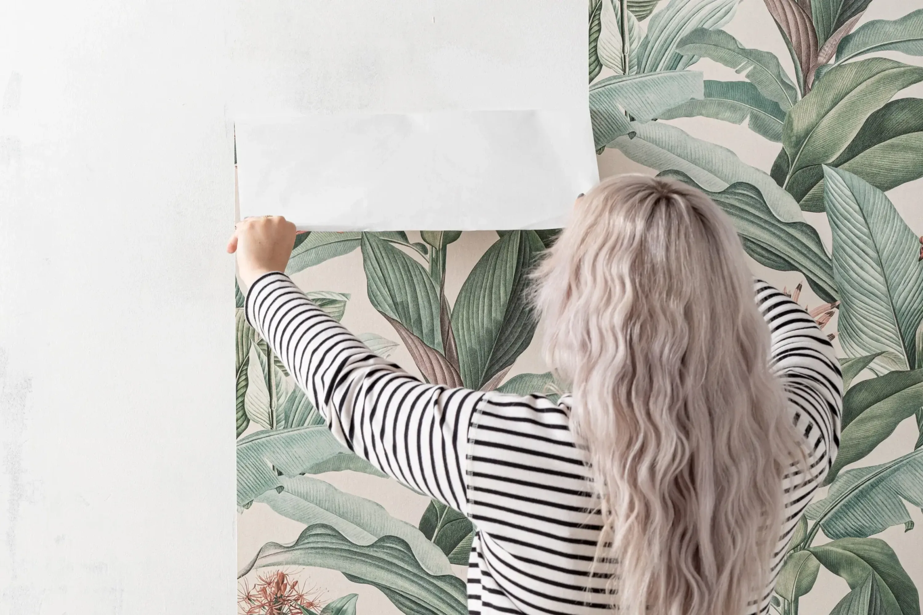 A woman with long, wavy blonde hair removes floral wallpaper from a white wall.