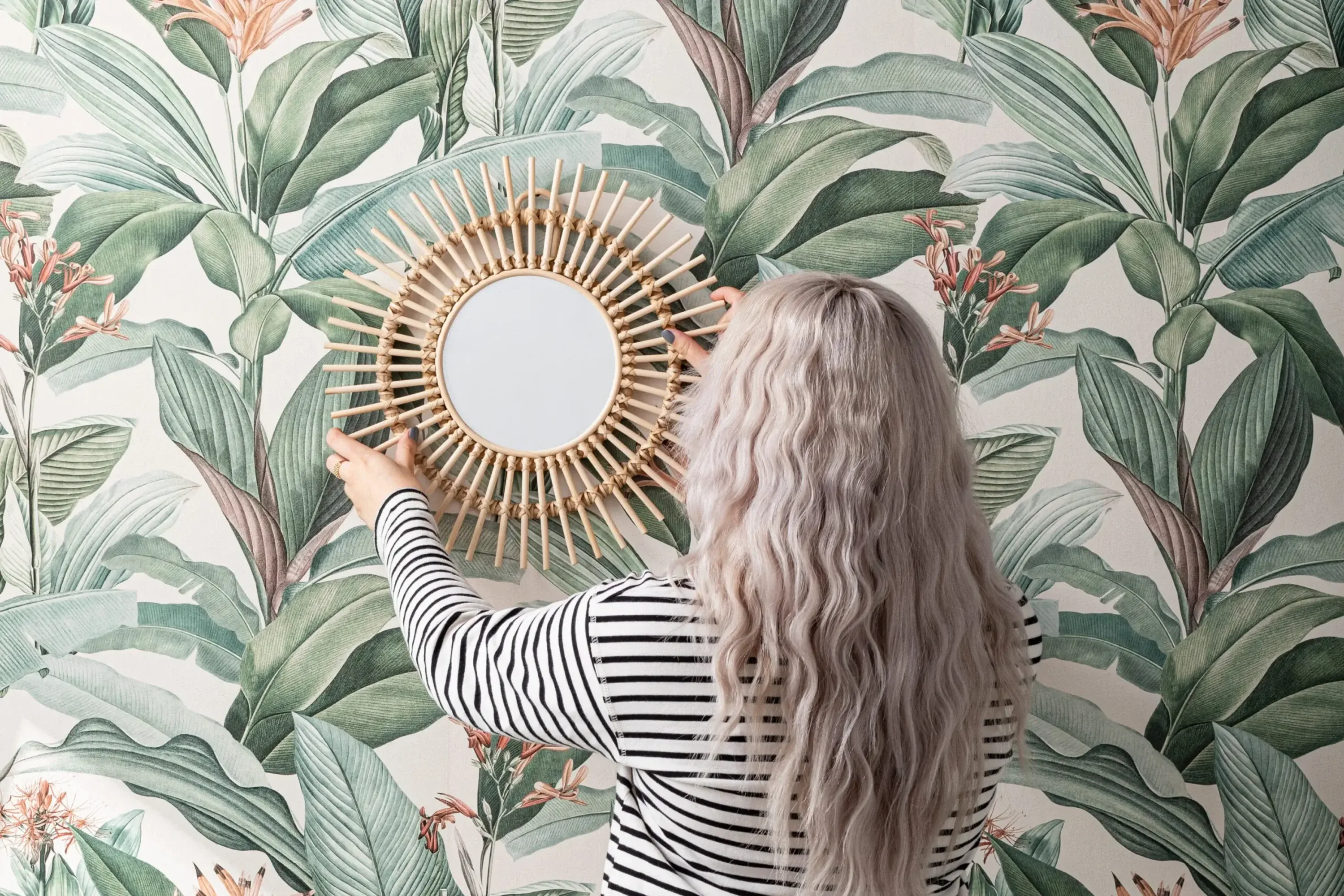 A woman hangs a round wicker mirror on a wall with green and pink botanical wallpaper.