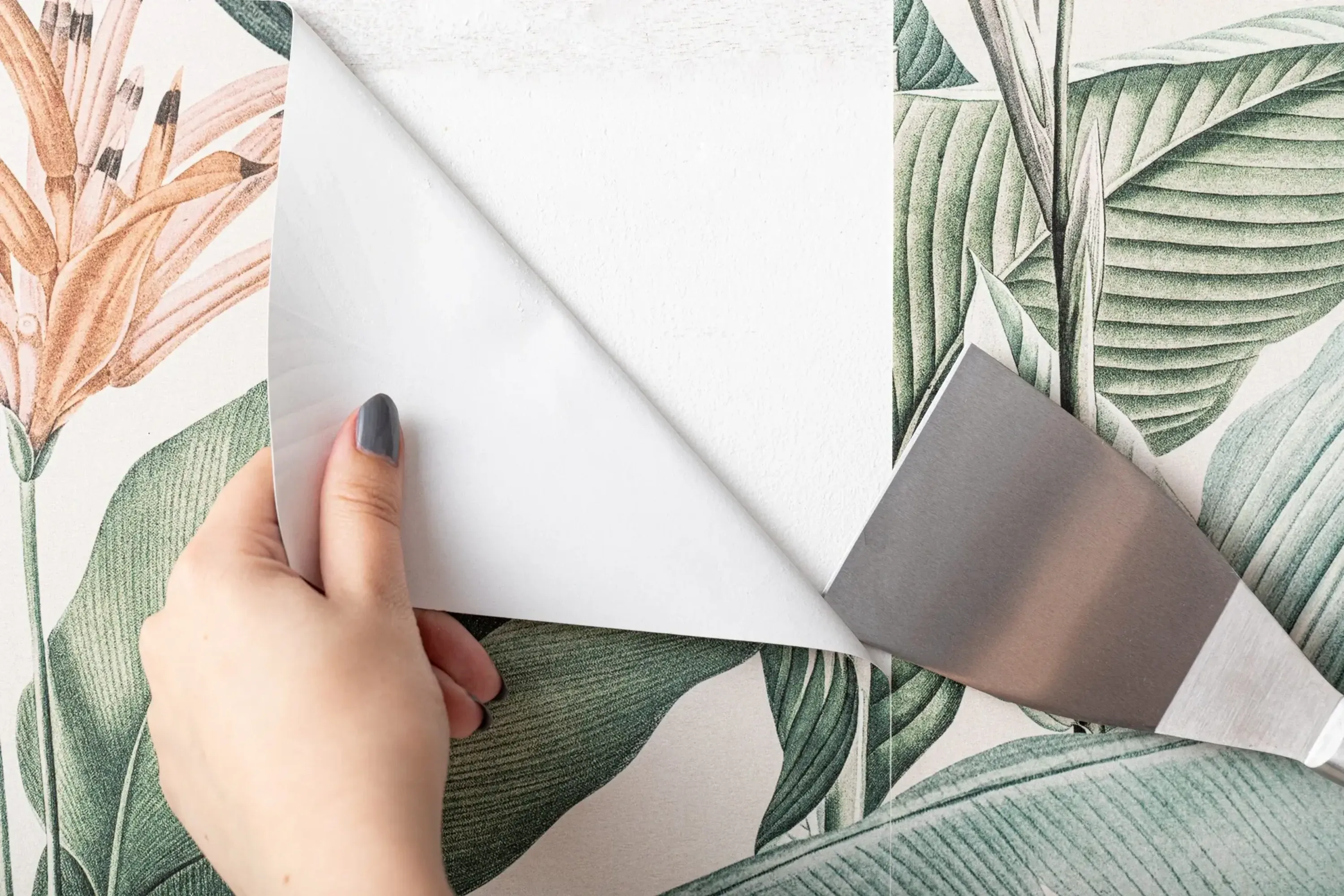 A person peels off botanical wallpaper from a white wall using a scraper.
