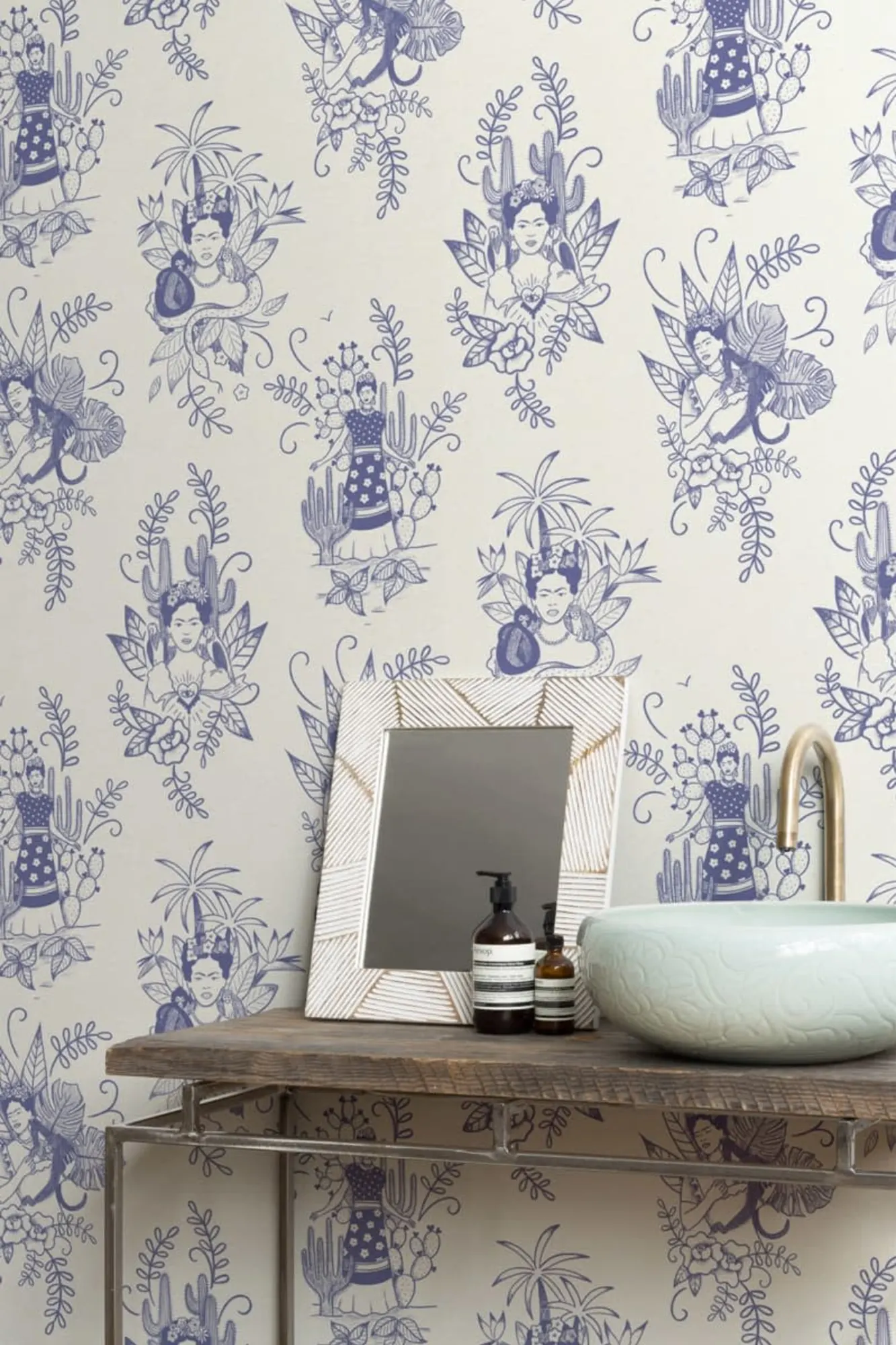 A bathroom with wallpaper featuring blue illustrations of Frida Kahlo among plants, a wooden vanity with a decorative mirror, and a ceramic vessel sink.