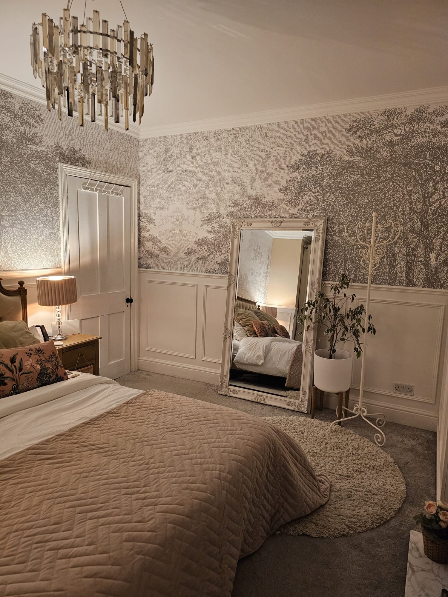 A bedroom with a chandelier, nature-themed wallpaper, a large standing mirror, a beige quilted bedspread, and a potted plant.