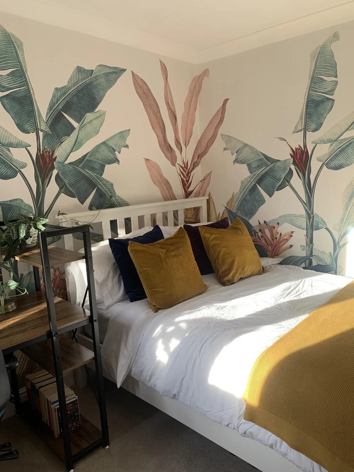 A bedroom with tropical leaf wallpaper, a white bed frame, mustard and navy throw pillows, and a wooden shelf with books and plants.
