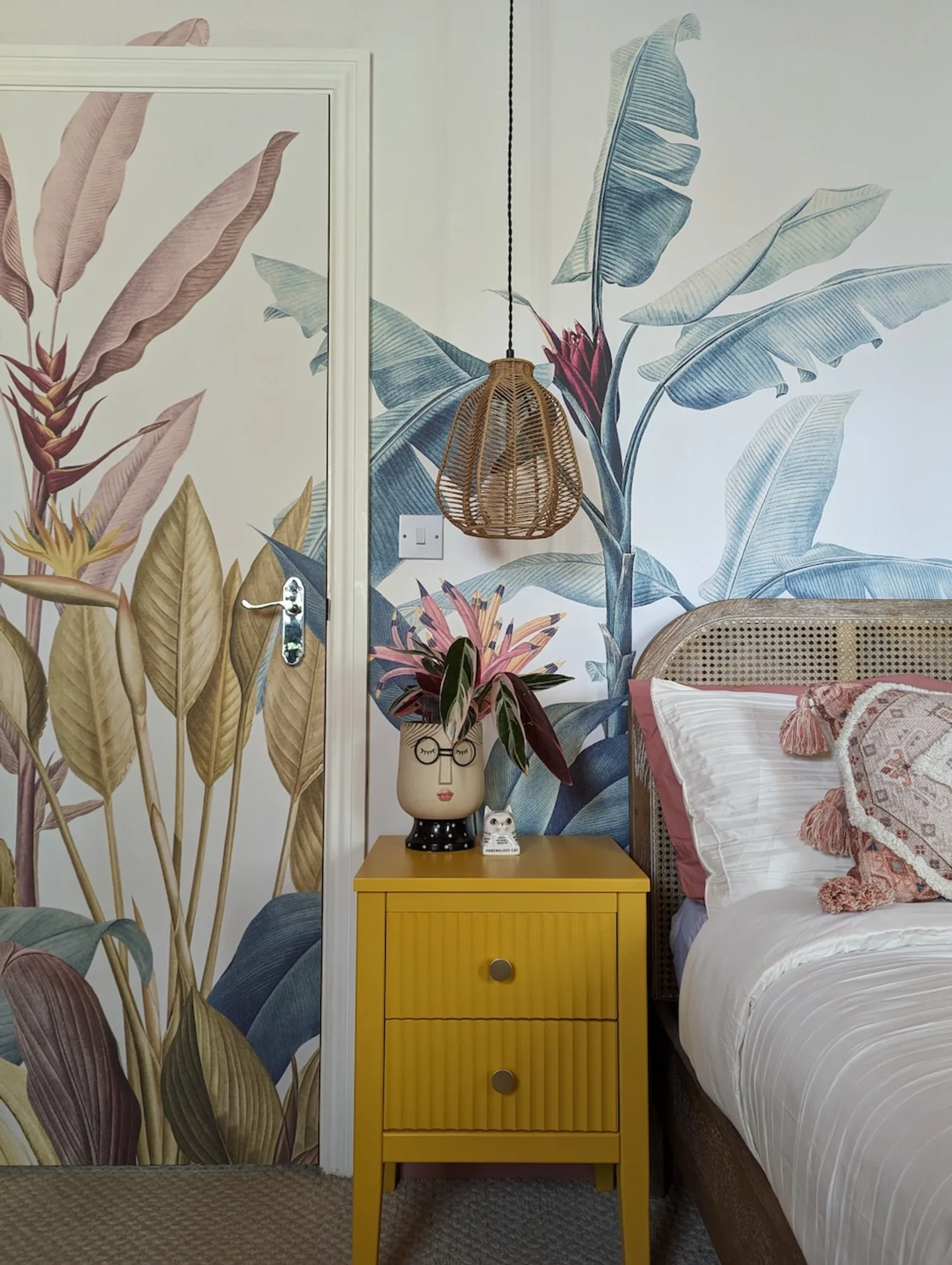 A bedroom with a bold, botanical-themed mural on the wall and door, a yellow nightstand, woven hanging lamp, and modern decor accents.