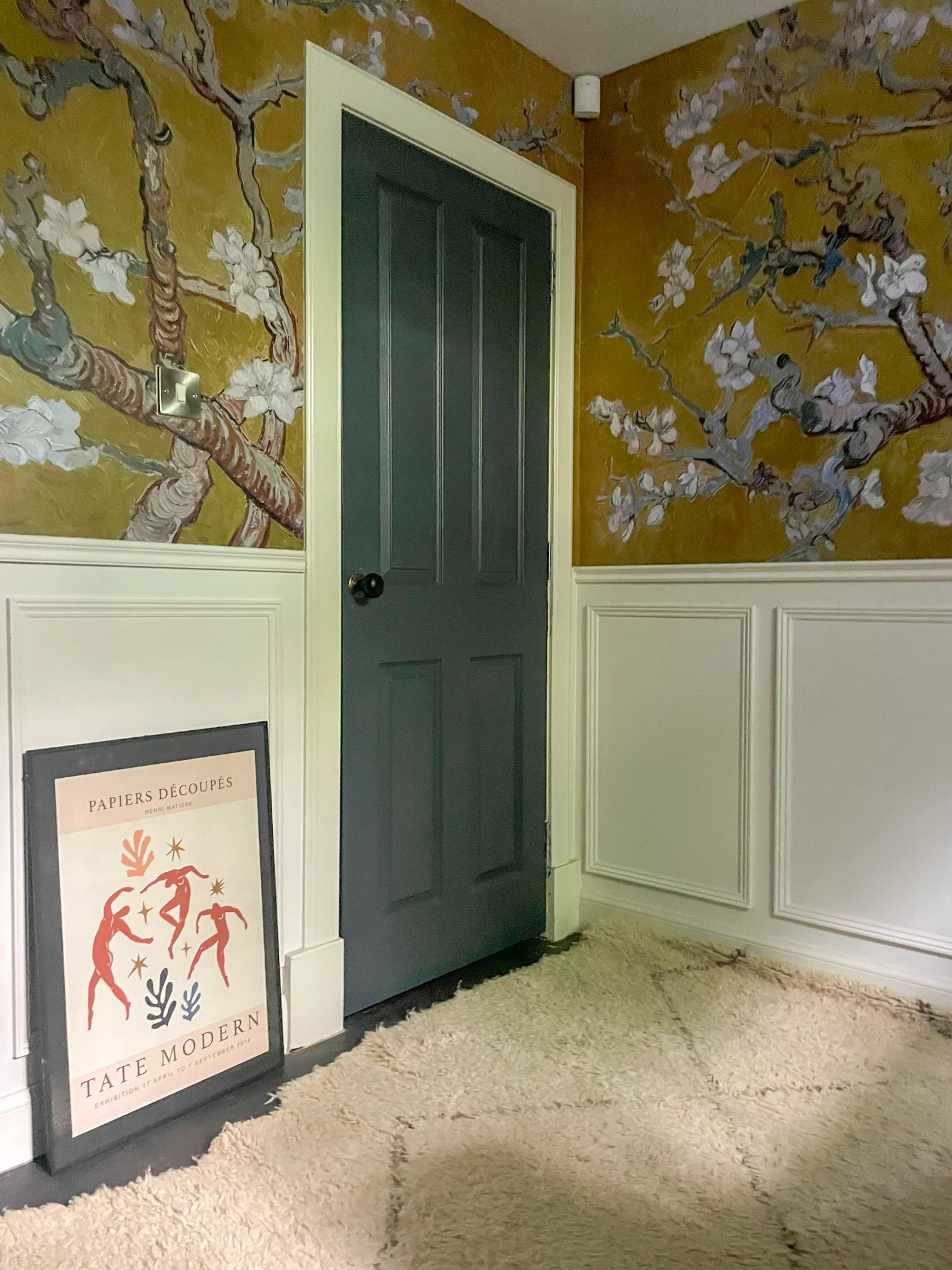 A room corner with floral wallpaper in yellow tones around a dark green door with a beige textured rug, and a framed Tate Modern poster leaning against the wall.
