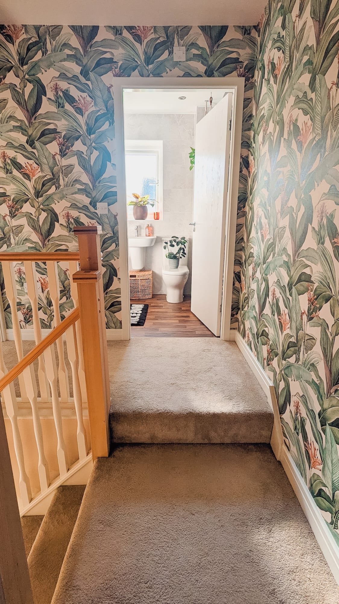 The image shows a hallway with tropical themed wallpaper decorating the walls around an open door to a small bathroom with wood flooring.